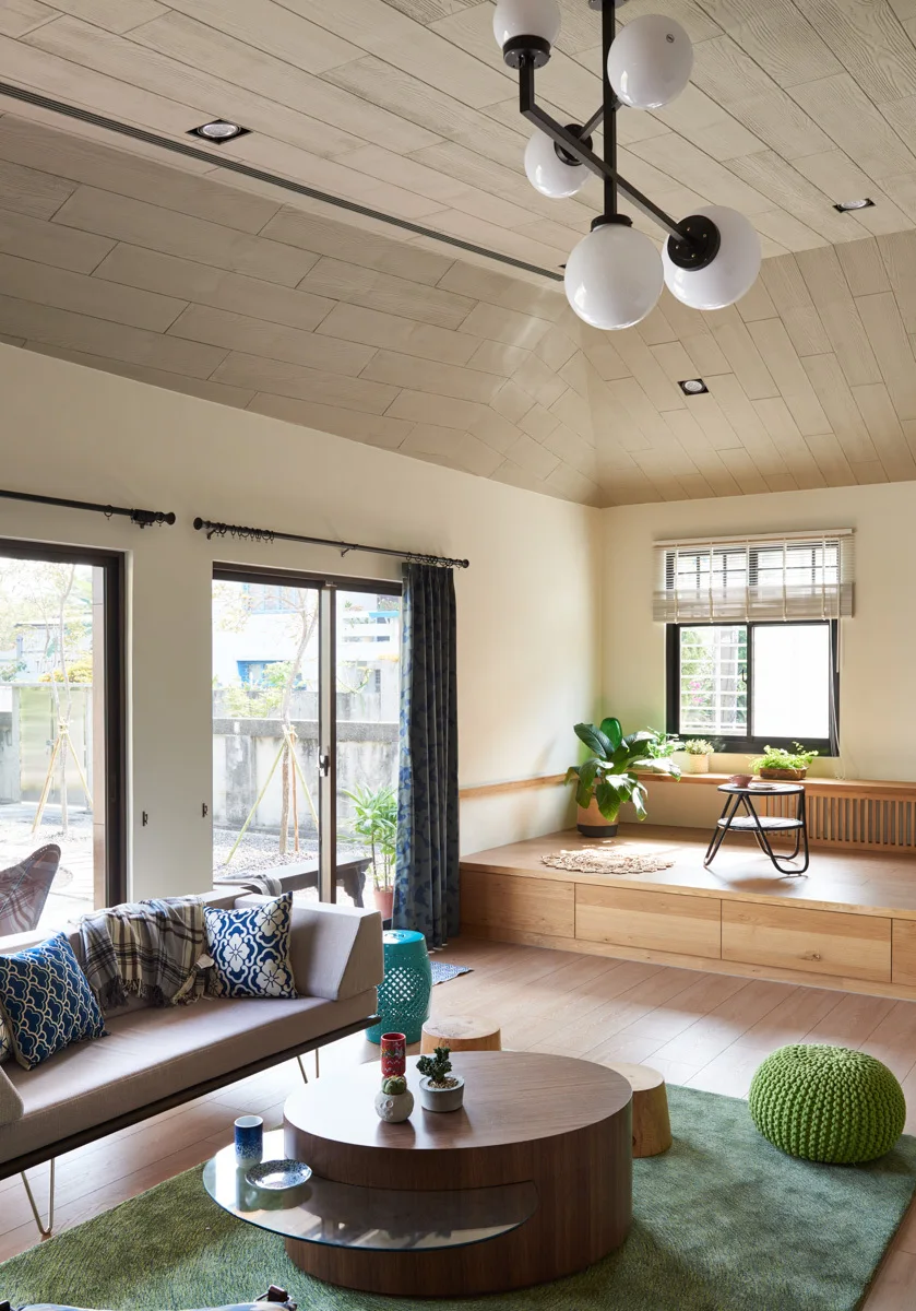A modern living room features a sloped wood-paneled ceiling, a raised wooden platform with storage drawers, a grey sofa, and a green area rug.