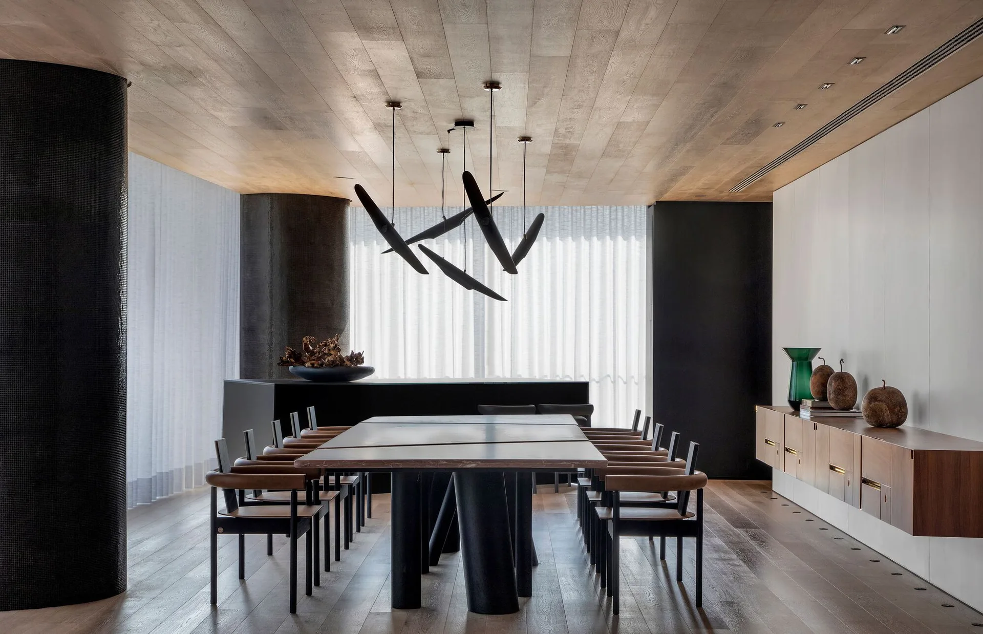 A spacious dining room features a long dark table, leather chairs, a wood-paneled ceiling, and sculptural black pendant lights in front of sheer drapes.