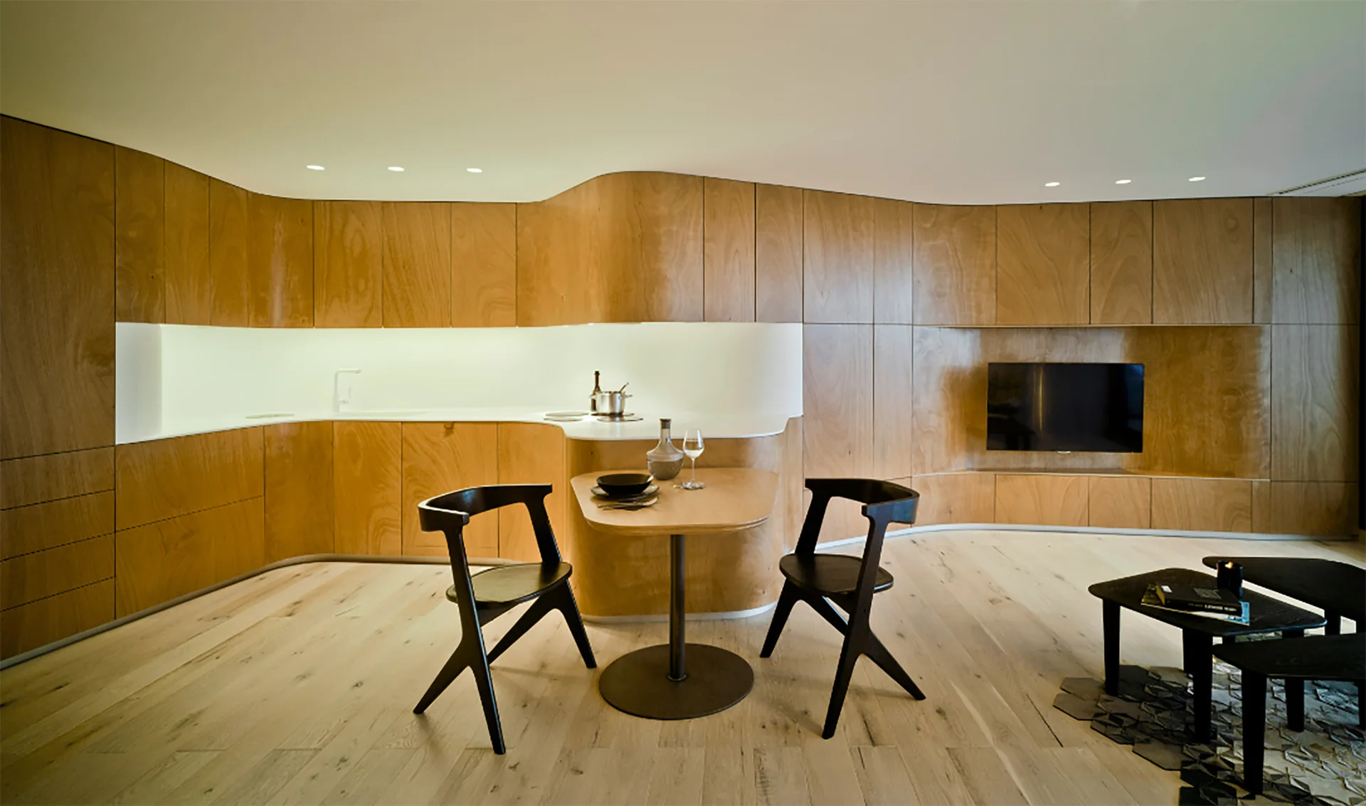 A modern open-plan kitchen and dining area featuring a continuous curved plywood wall, a white countertop, and a dark wooden table with two chairs.
