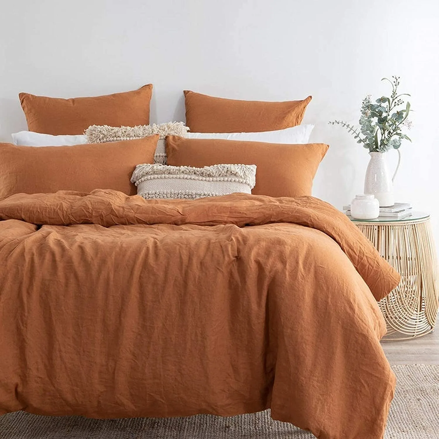 A bed with terracotta orange linen sheets and textured cream pillows sits next to a round wicker nightstand holding a white vase with greenery.