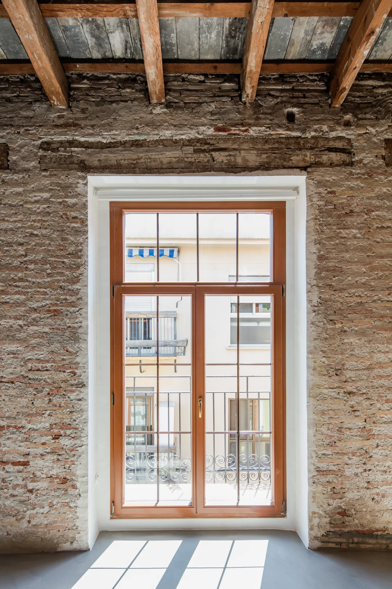 A wooden gridded window is centered in a rustic exposed brick wall beneath rough-hewn ceiling beams.