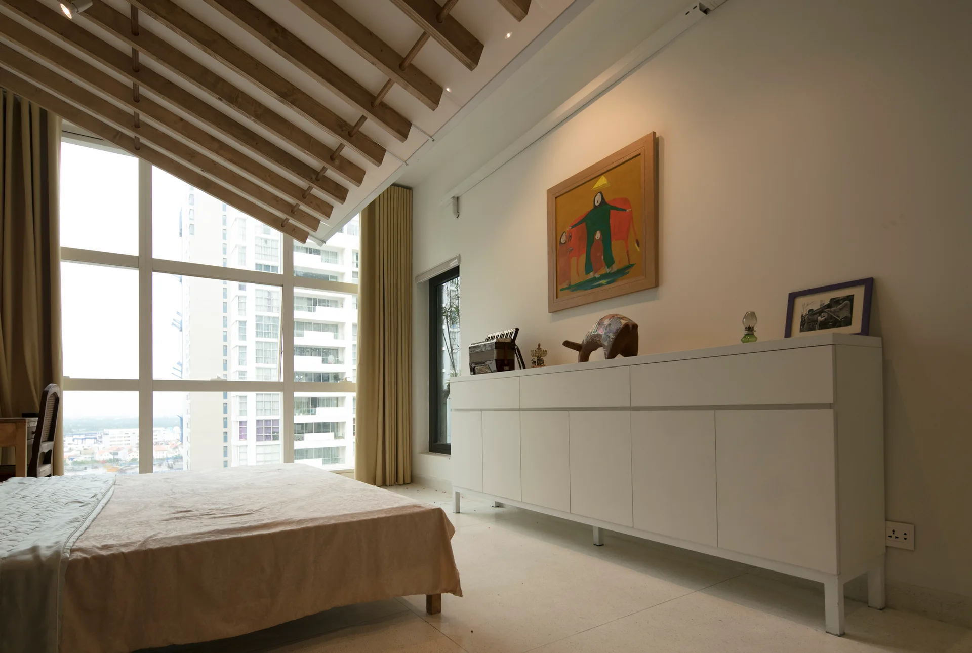 A low bed and a long white credenza sit in a modern bedroom featuring an angled ceiling with exposed wooden rafters next to large windows.