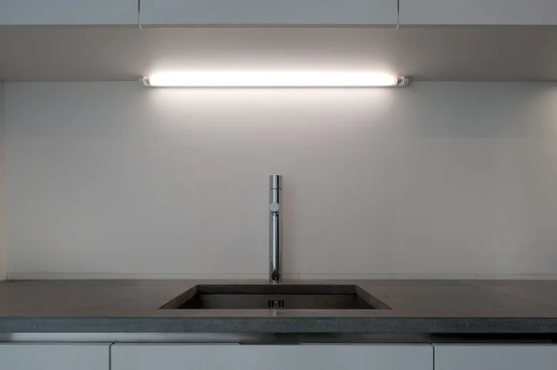 A minimalist kitchen sink with a tall chrome faucet is set into a grey countertop under a bright linear task light.