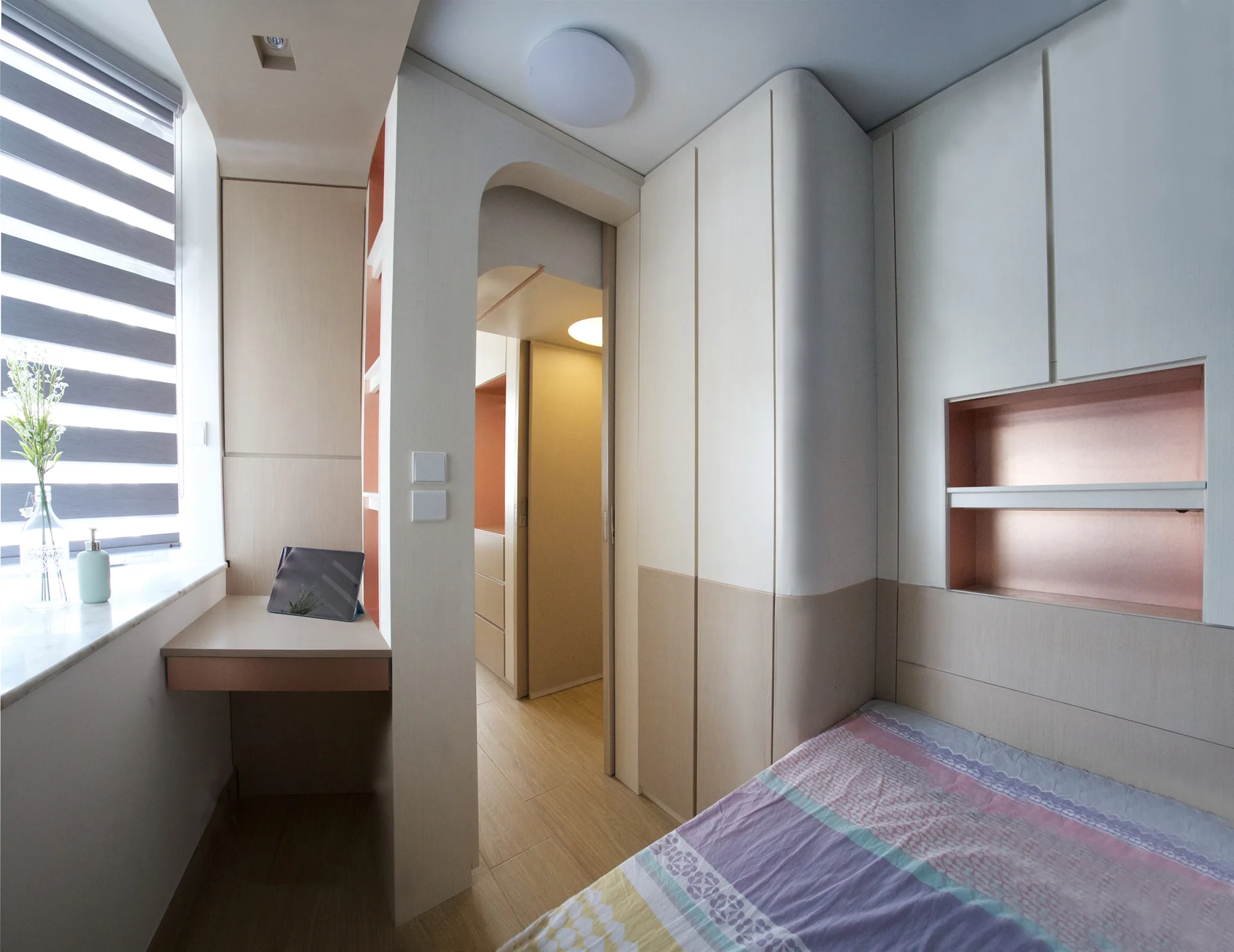 A compact modern bedroom features beige built-in cabinetry, a work desk by a window, and a bed with a recessed copper-colored shelf.