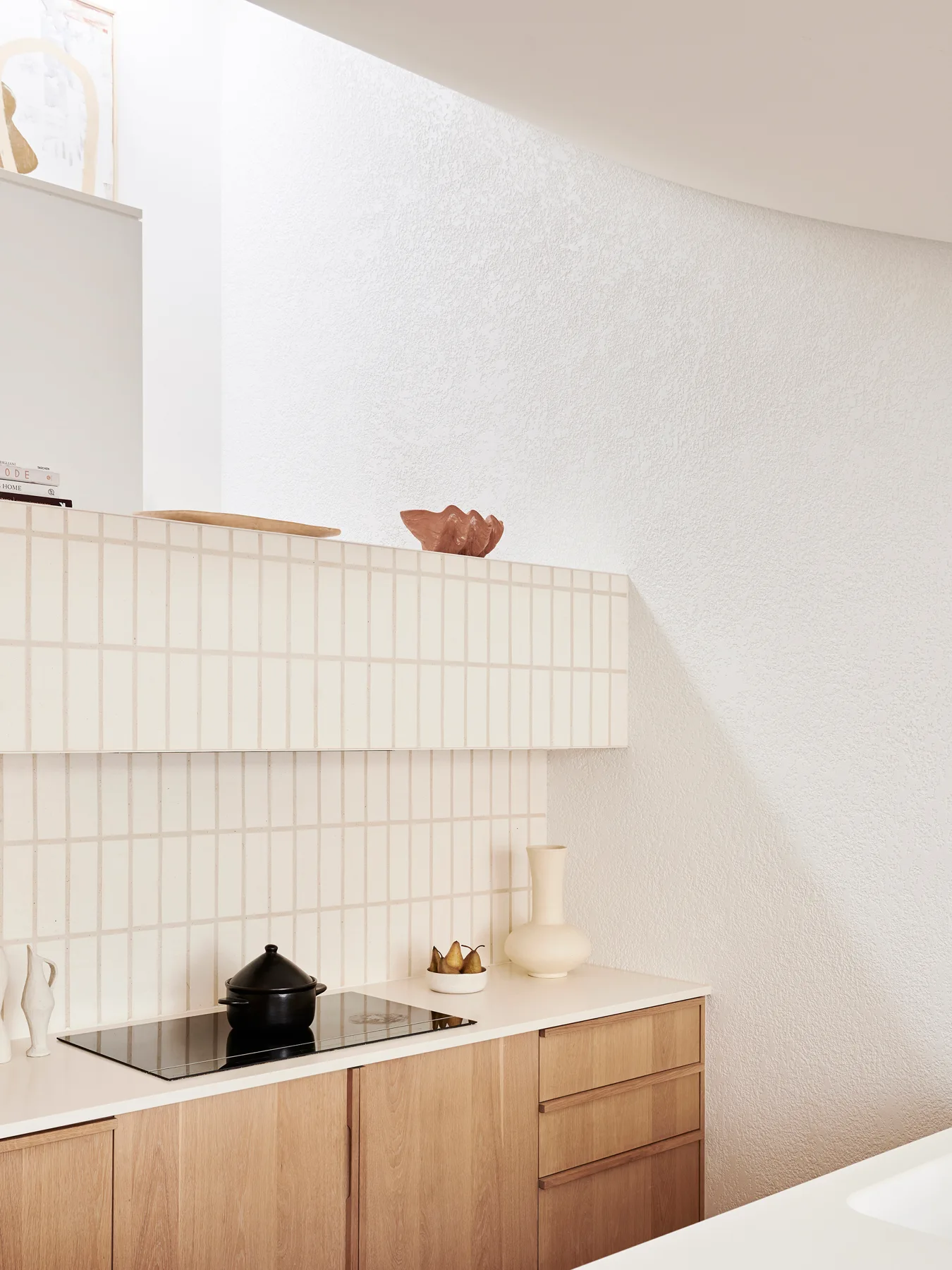 A minimalist kitchen featuring light wood lower cabinets, an induction cooktop, and a white grid-tiled backsplash that forms a built-in ledge.