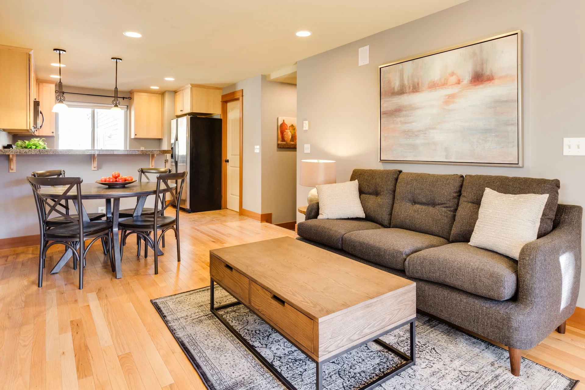 A transitional living room features a grey sofa, a wooden coffee table, a patterned rug, and a dining table with chairs in the background.