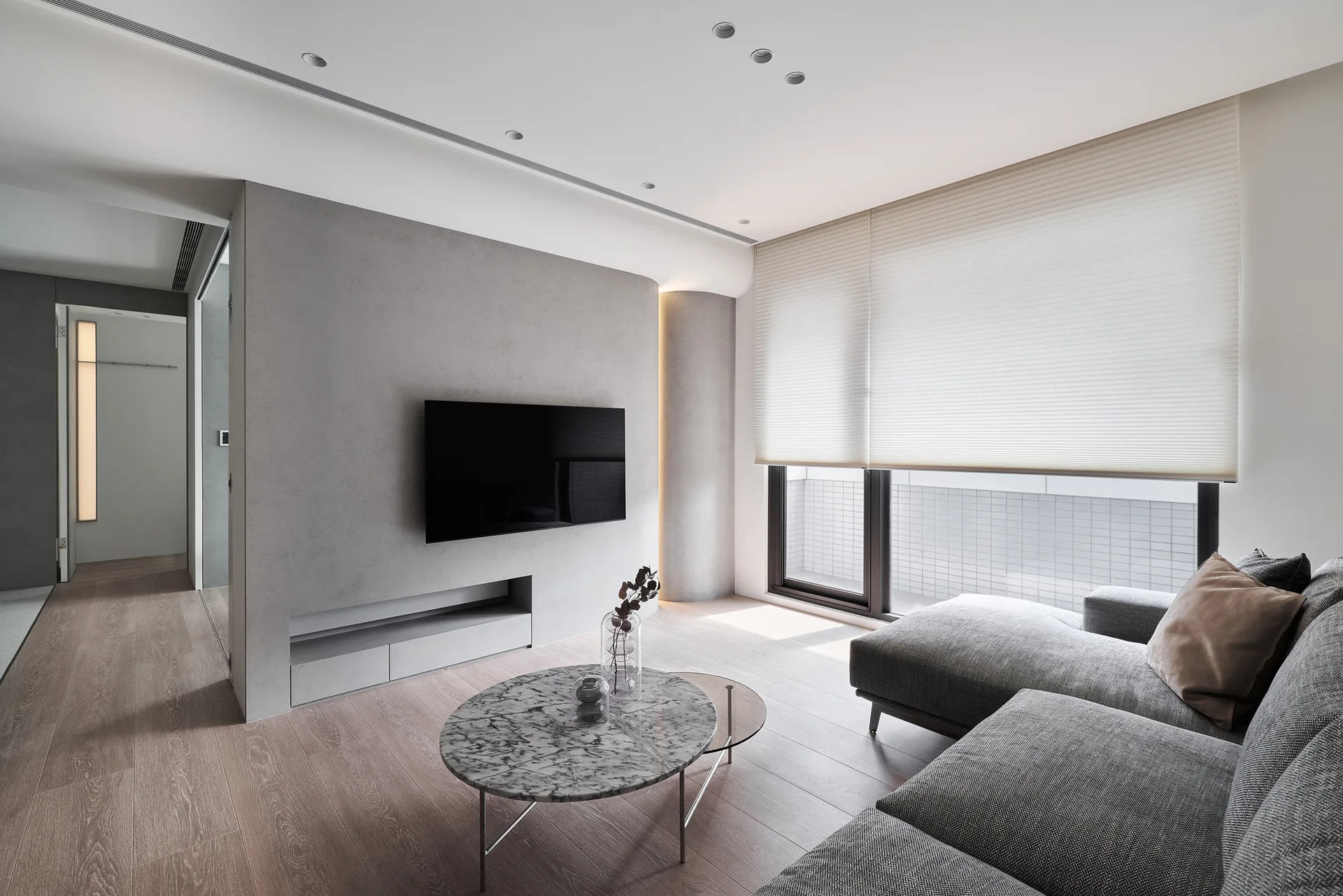 A modern living room features a grey sectional sofa, a round marble coffee table, and a wall-mounted TV on a grey partition wall.