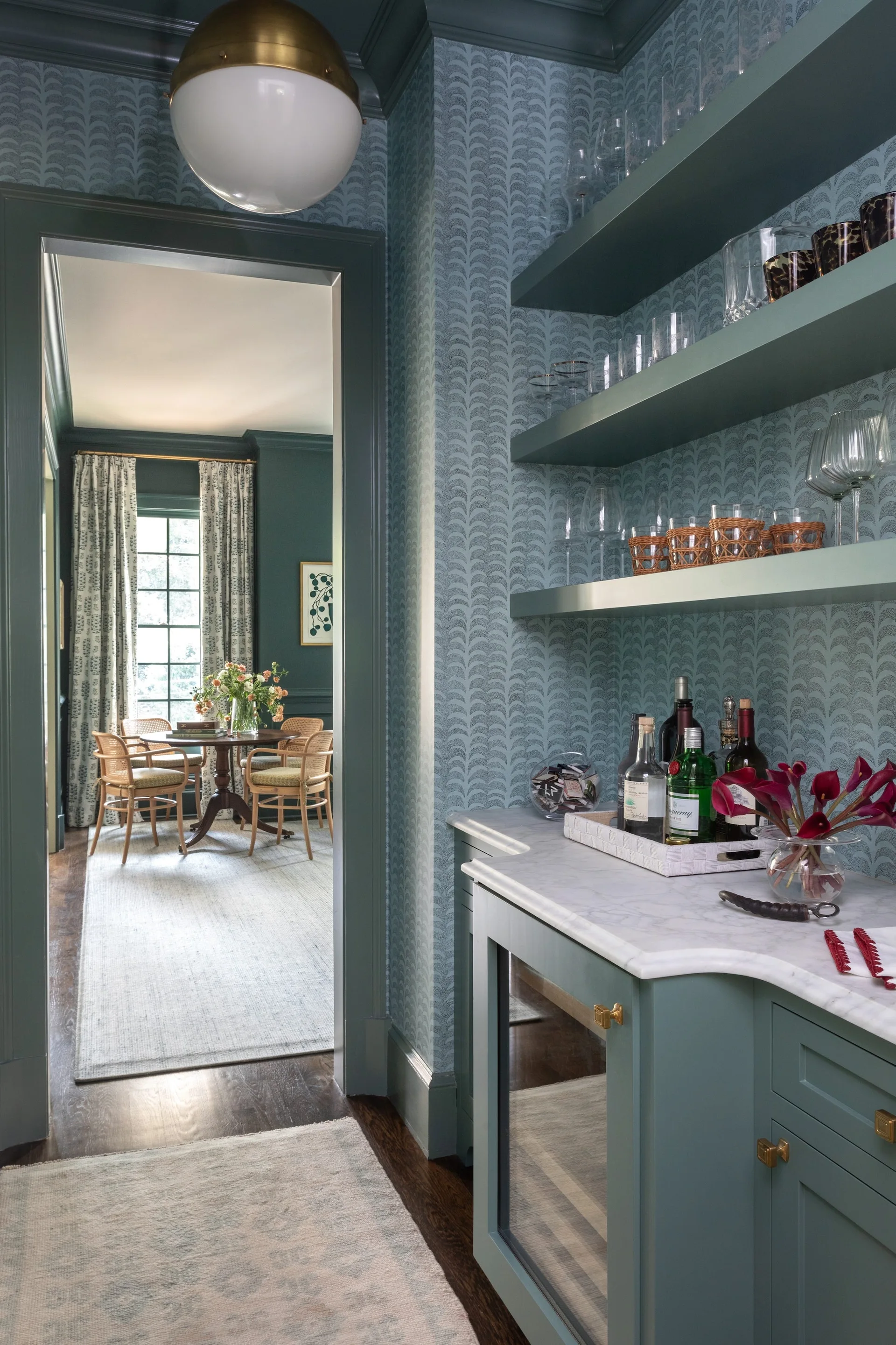 A built-in blue-green home bar with open shelves, a marble countertop, and patterned wallpaper looks through a doorway into a dining room.