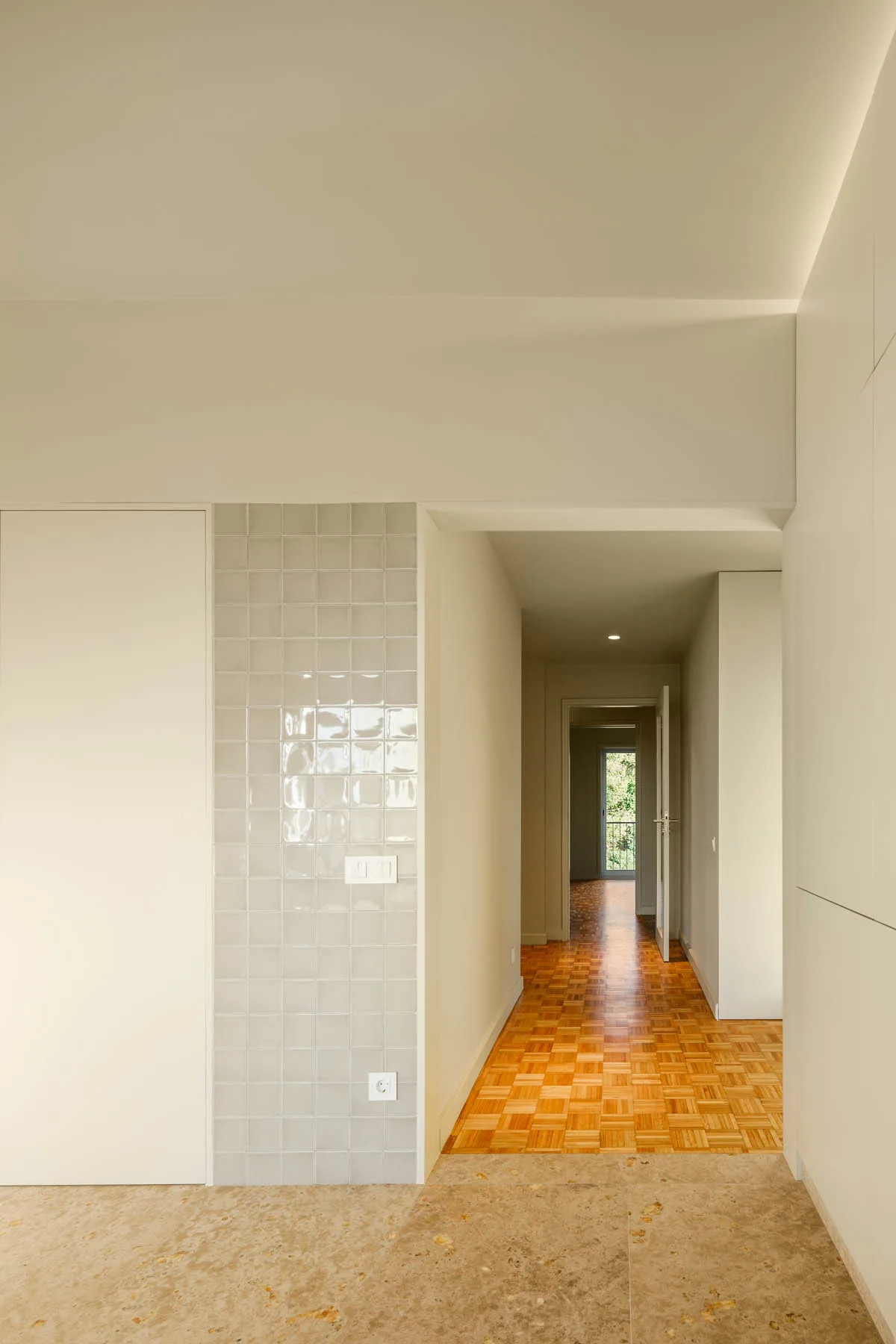 A minimalist hallway featuring white built-in storage, a glossy grey-tiled wall panel, and warm parquet wood flooring.