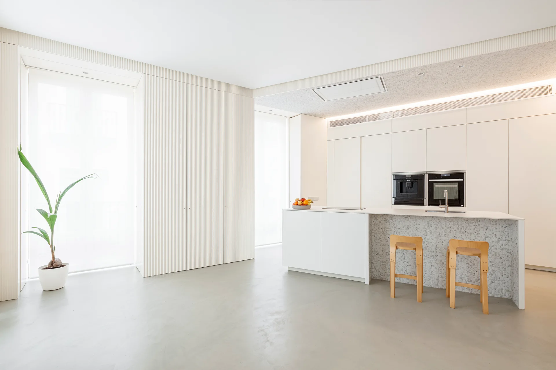 A minimalist white kitchen features a mosaic tile island with two wooden stools, fluted wall panels, and a seamless grey concrete floor.