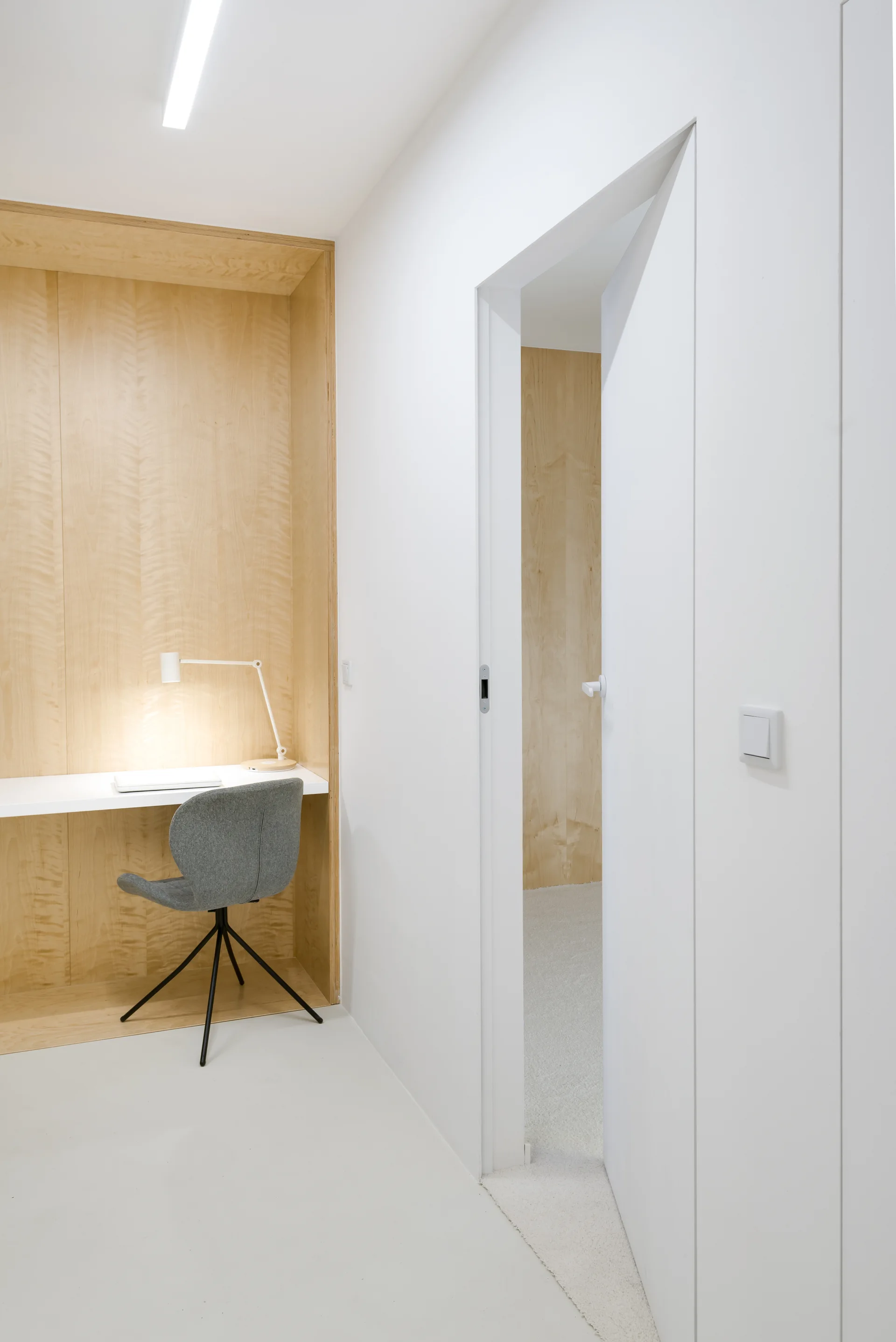 A minimalist built-in plywood desk nook with a white floating desktop, a grey office chair, and an adjacent open doorway in a white room.