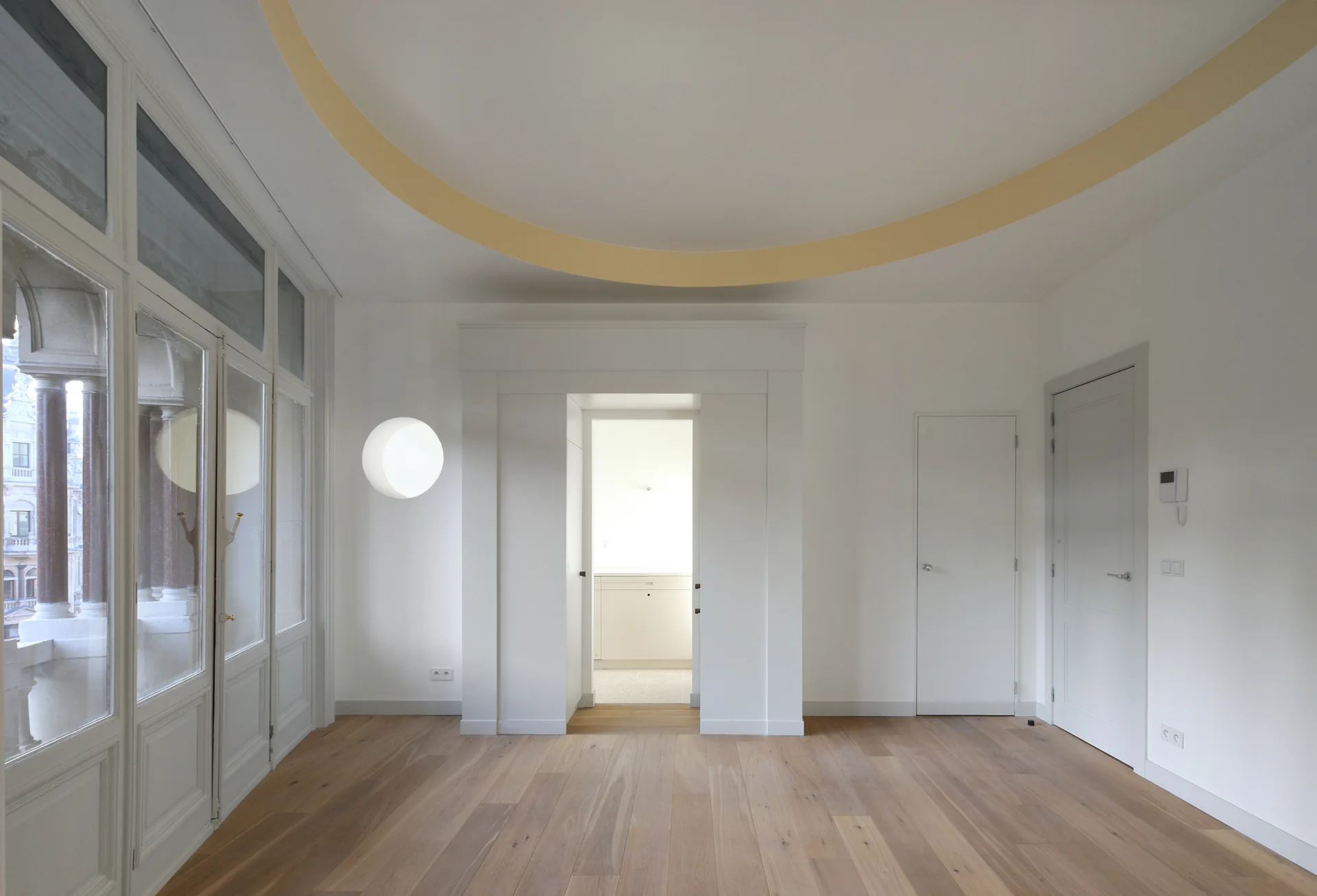 A minimalist empty room features a yellow painted ring on the ceiling, light hardwood floors, and a wall of glass-paned doors.