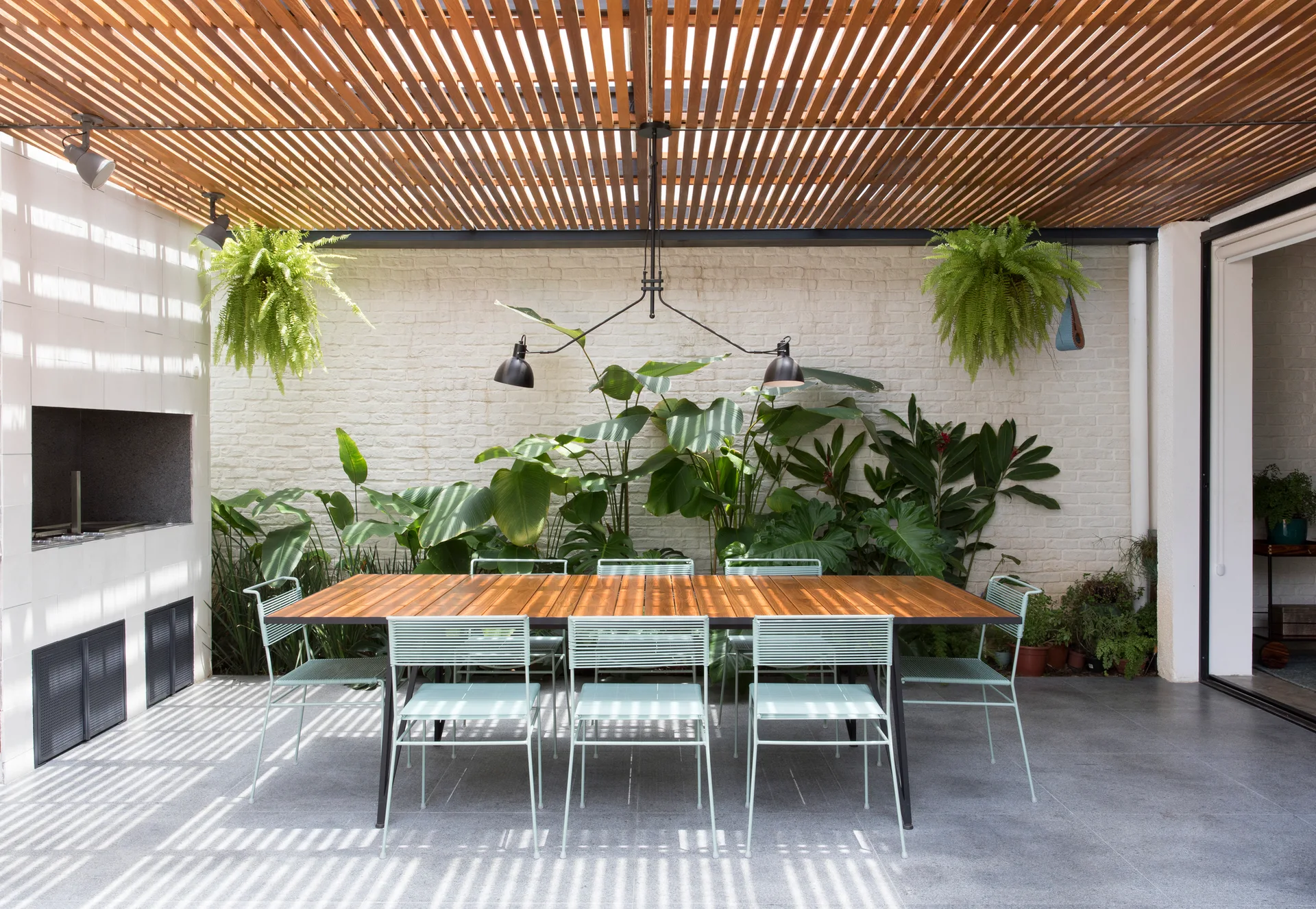 A modern outdoor patio featuring a wooden dining table with light green chairs under a slatted pergola next to a built-in brick barbecue.