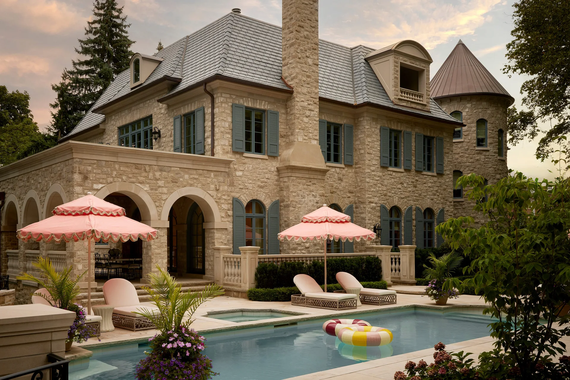 A large stone chateau-style home with blue window shutters features an in-ground swimming pool lined with lounge chairs and pink ruffled umbrellas.