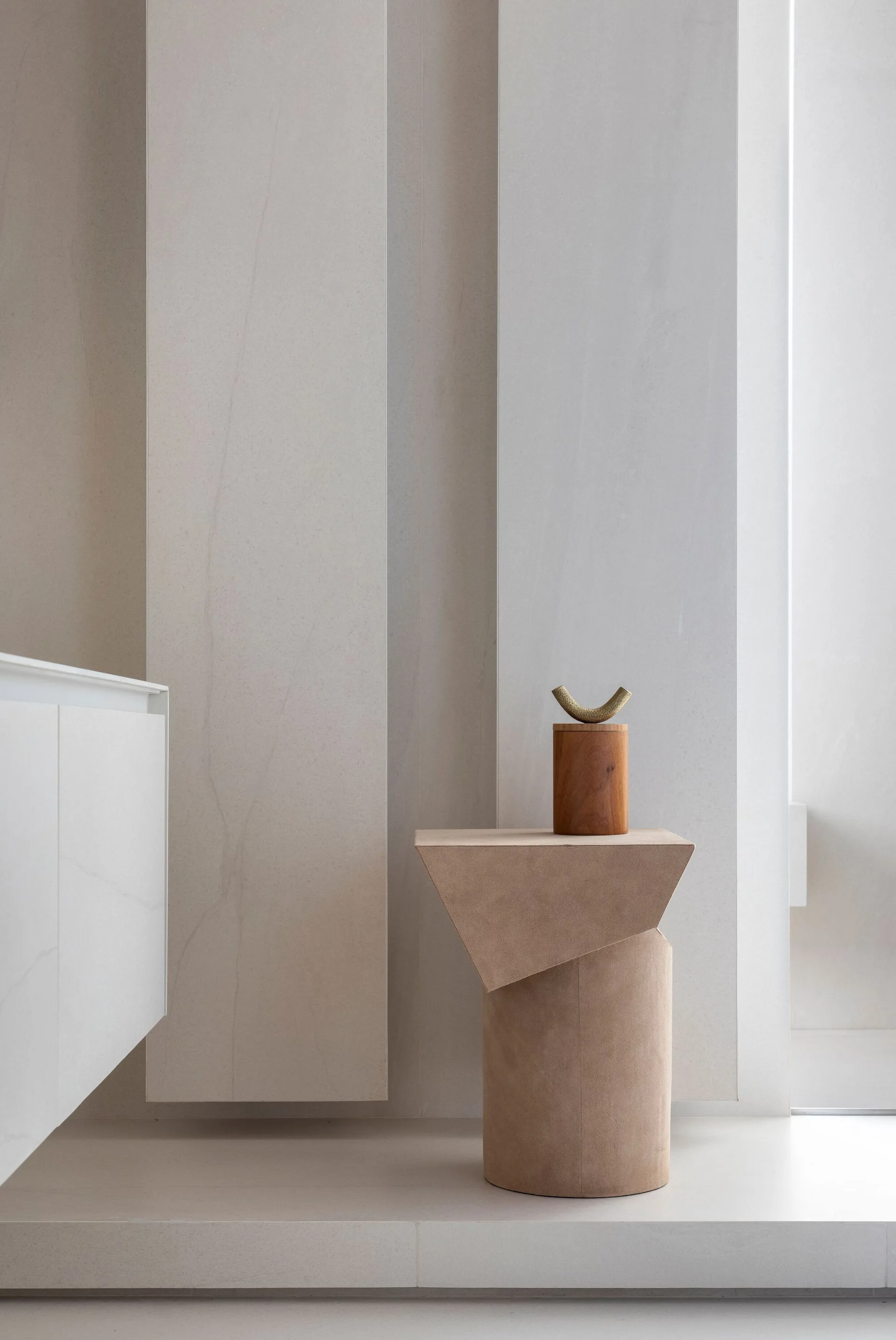 A sculptural beige side table stands against large vertical stone wall panels, holding a small wooden cylinder decorative object.