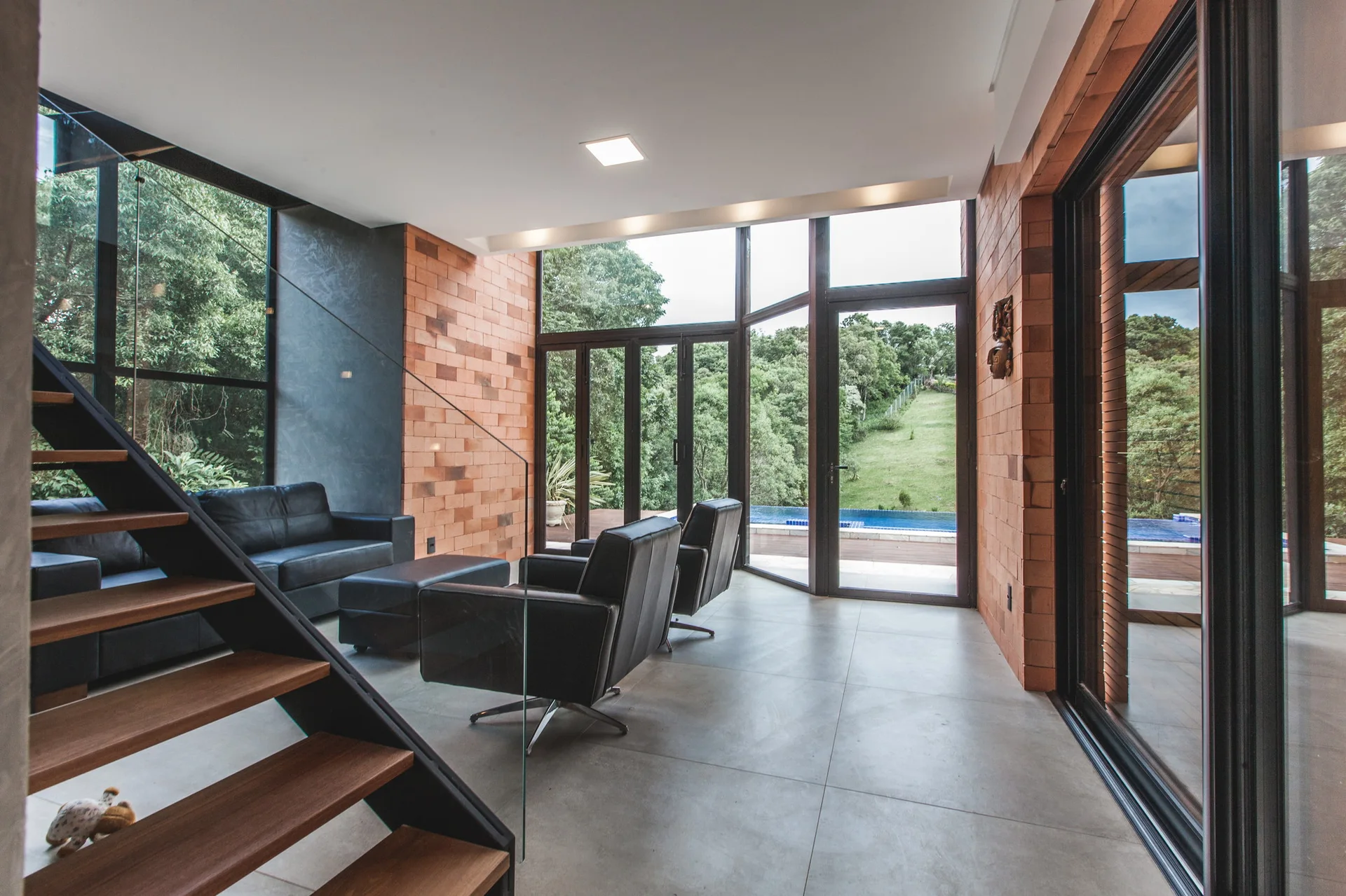 A modern living room features black leather seating, an exposed red brick wall, and a floating wooden staircase leading past floor-to-ceiling glass windows.