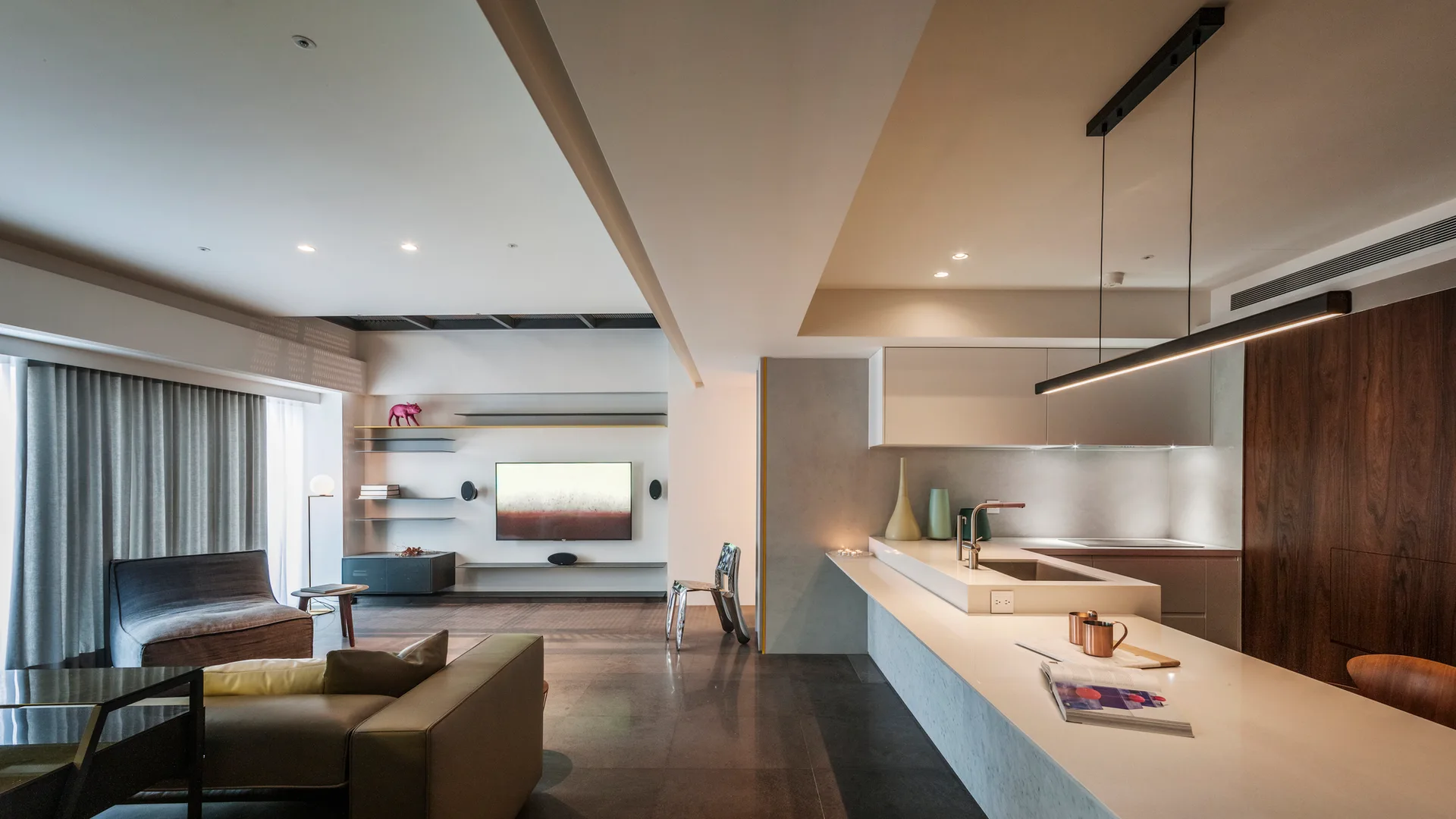 An open-concept living area features a long white kitchen island with a sink, a brown sofa, and a wall-mounted television with sleek floating shelves.