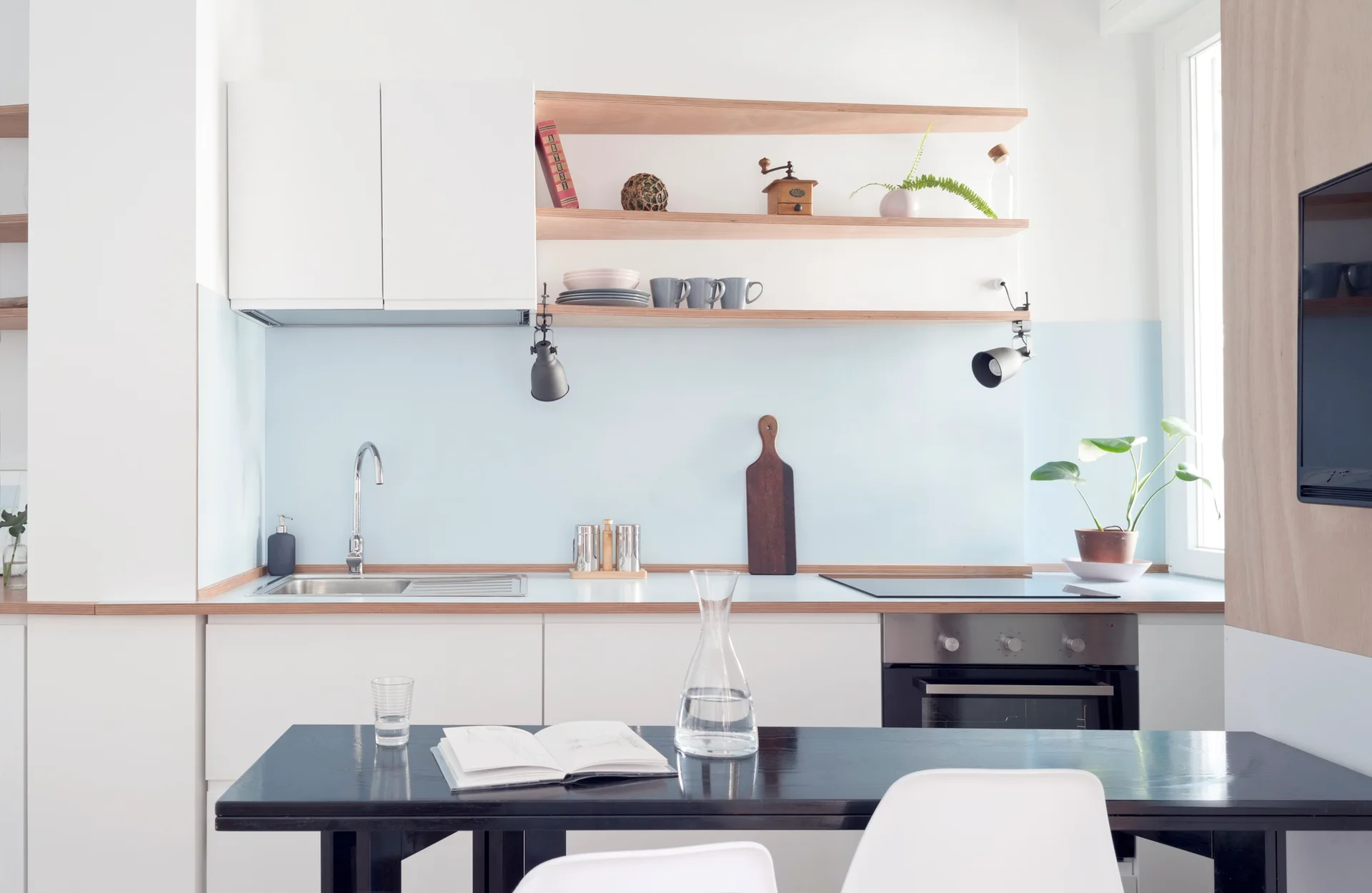 A modern Scandinavian kitchen features white cabinets, a pale blue backsplash, light wood open shelves, and a black dining table.