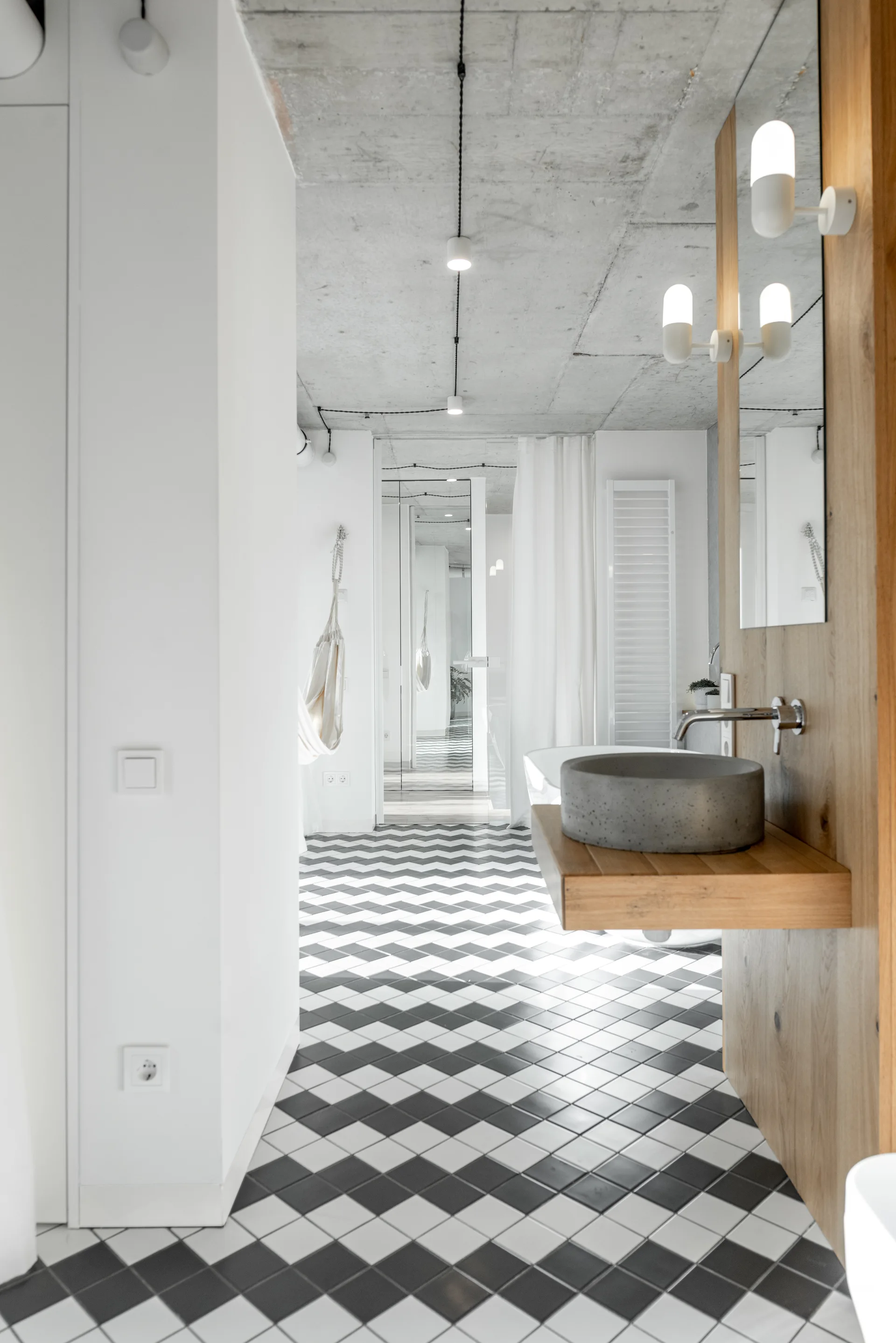A minimalist bathroom features a concrete vessel sink on a wood floating vanity over a black-and-white diamond tiled floor beneath a concrete ceiling.