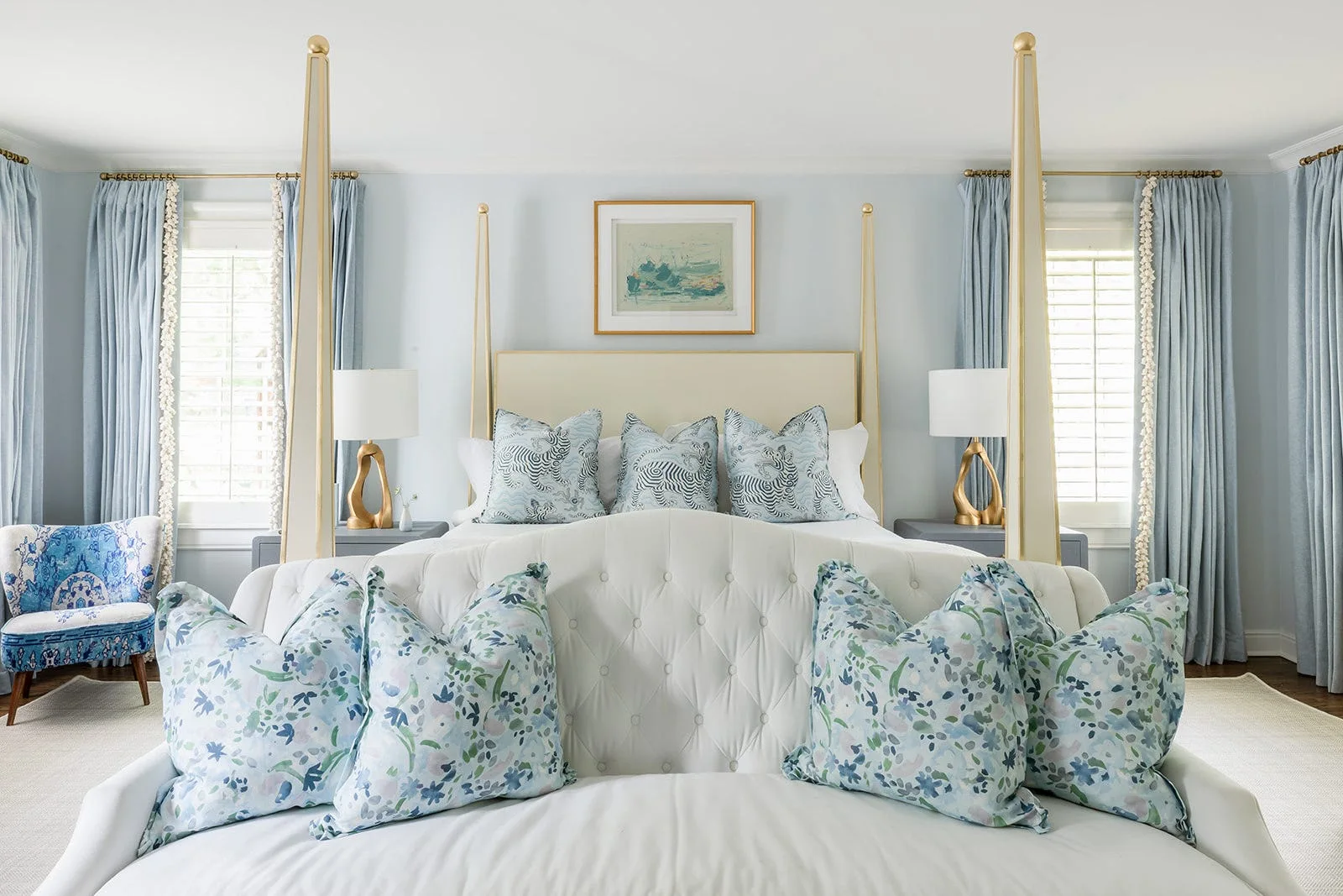 A bedroom features a gold four-poster bed with a tufted bench at the foot, decorated with patterned blue and white throw pillows.