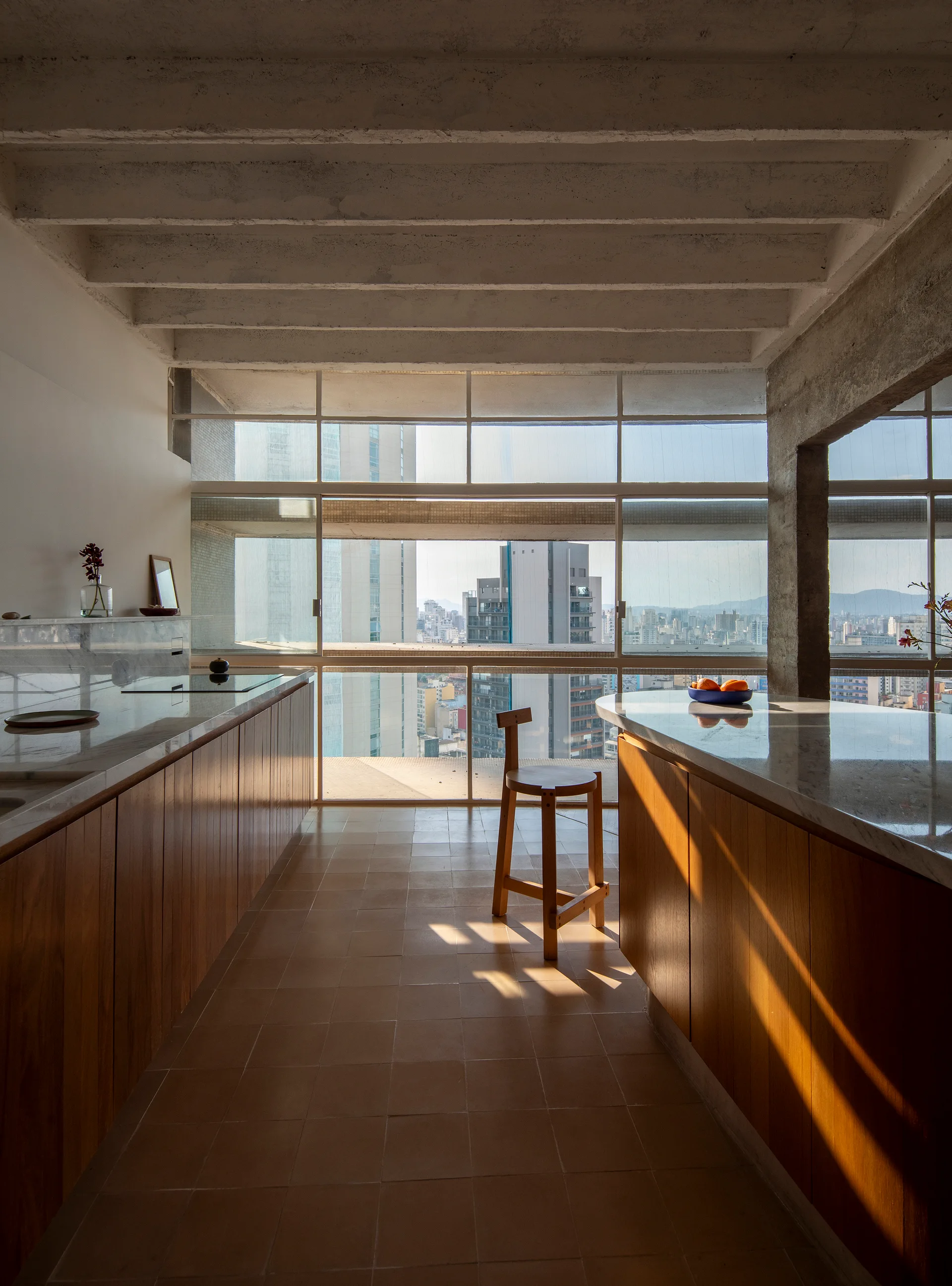 A modern apartment kitchen with warm wood-toned cabinets, stone countertops, an exposed concrete ceiling, and a large window overlooking a city skyline.