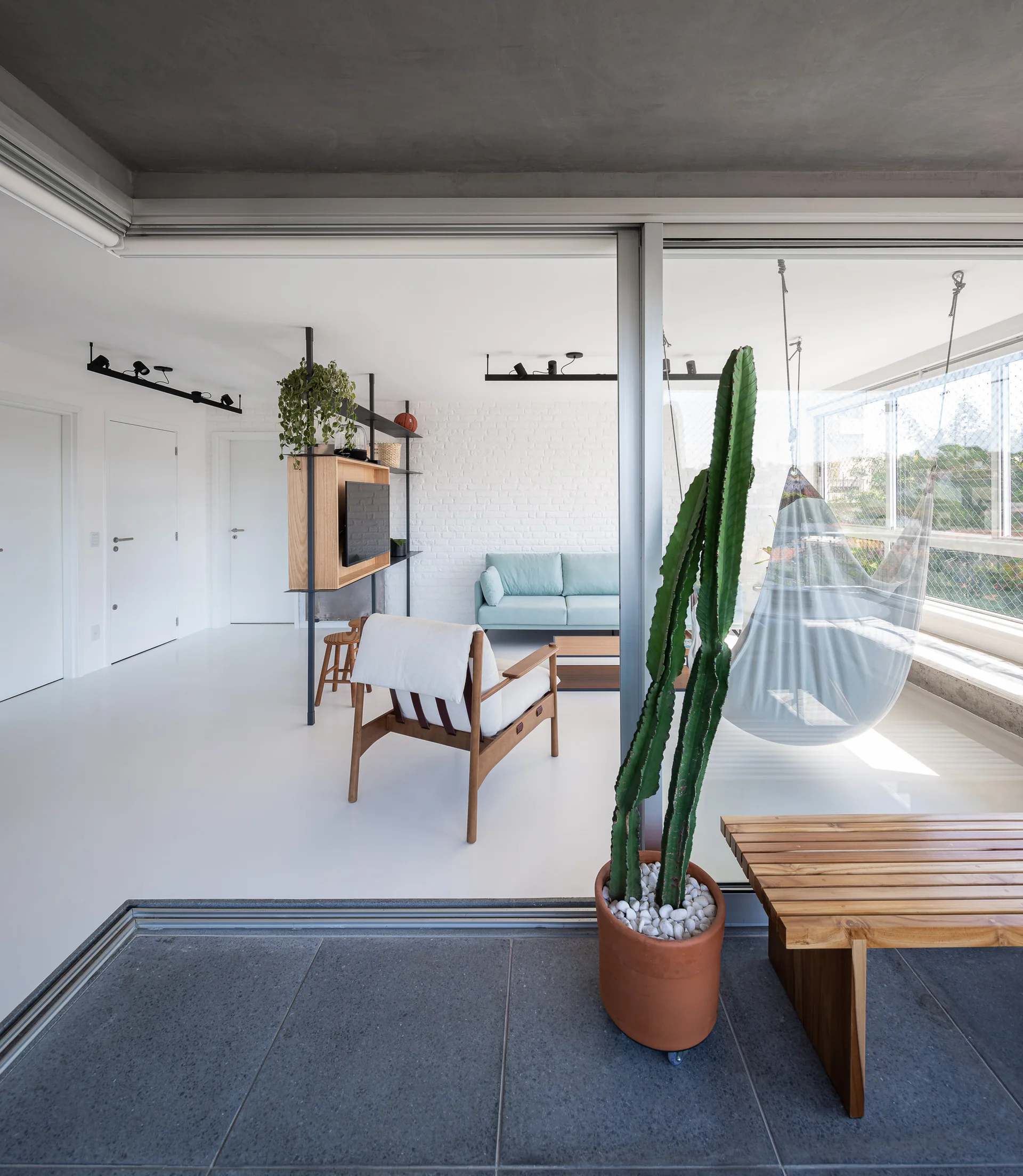 A minimalist living room with white floors features a light blue sofa, a wooden lounge chair, and a sliding glass door opening to a balcony with a hammock.