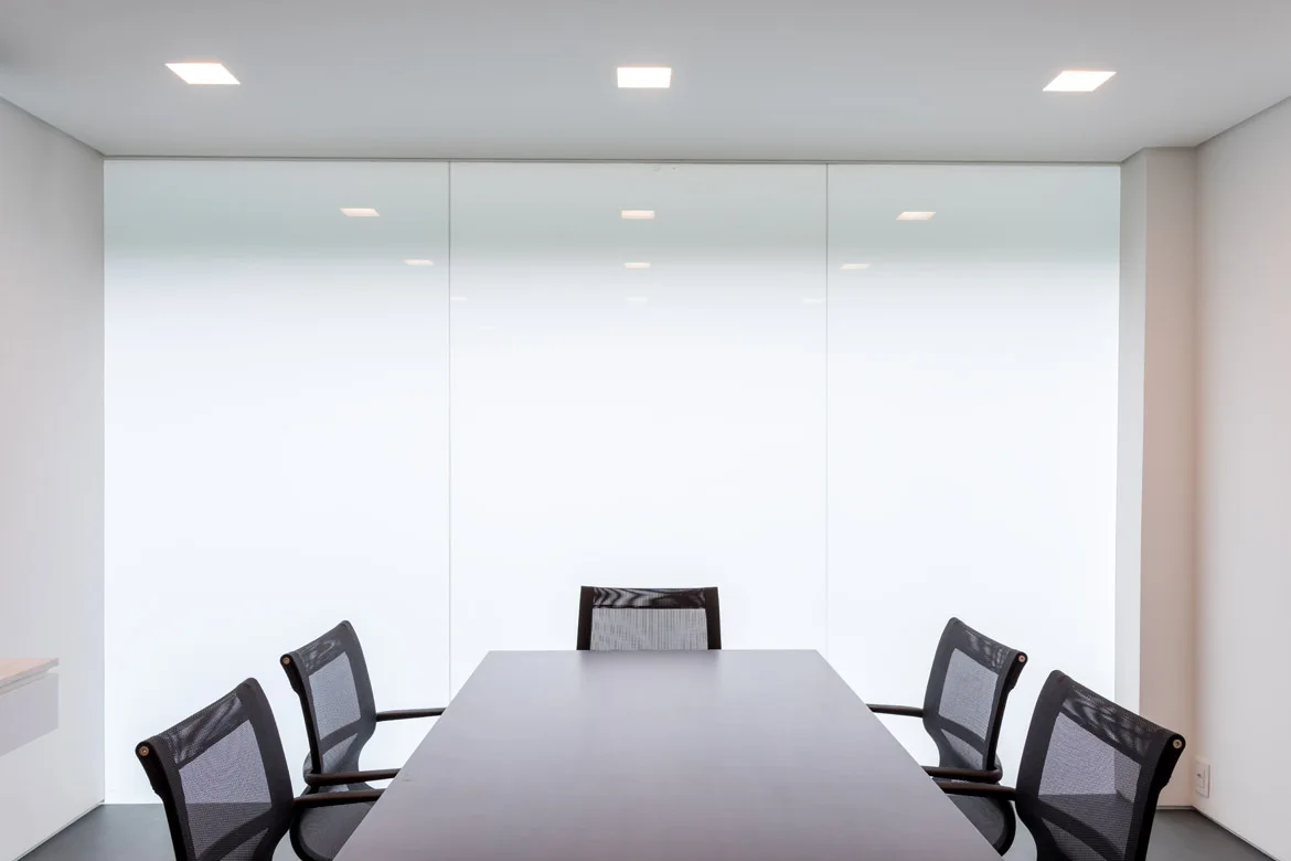 A dark rectangular conference table surrounded by black mesh office chairs sits before a large seamless white paneled wall under recessed square ceiling lights.