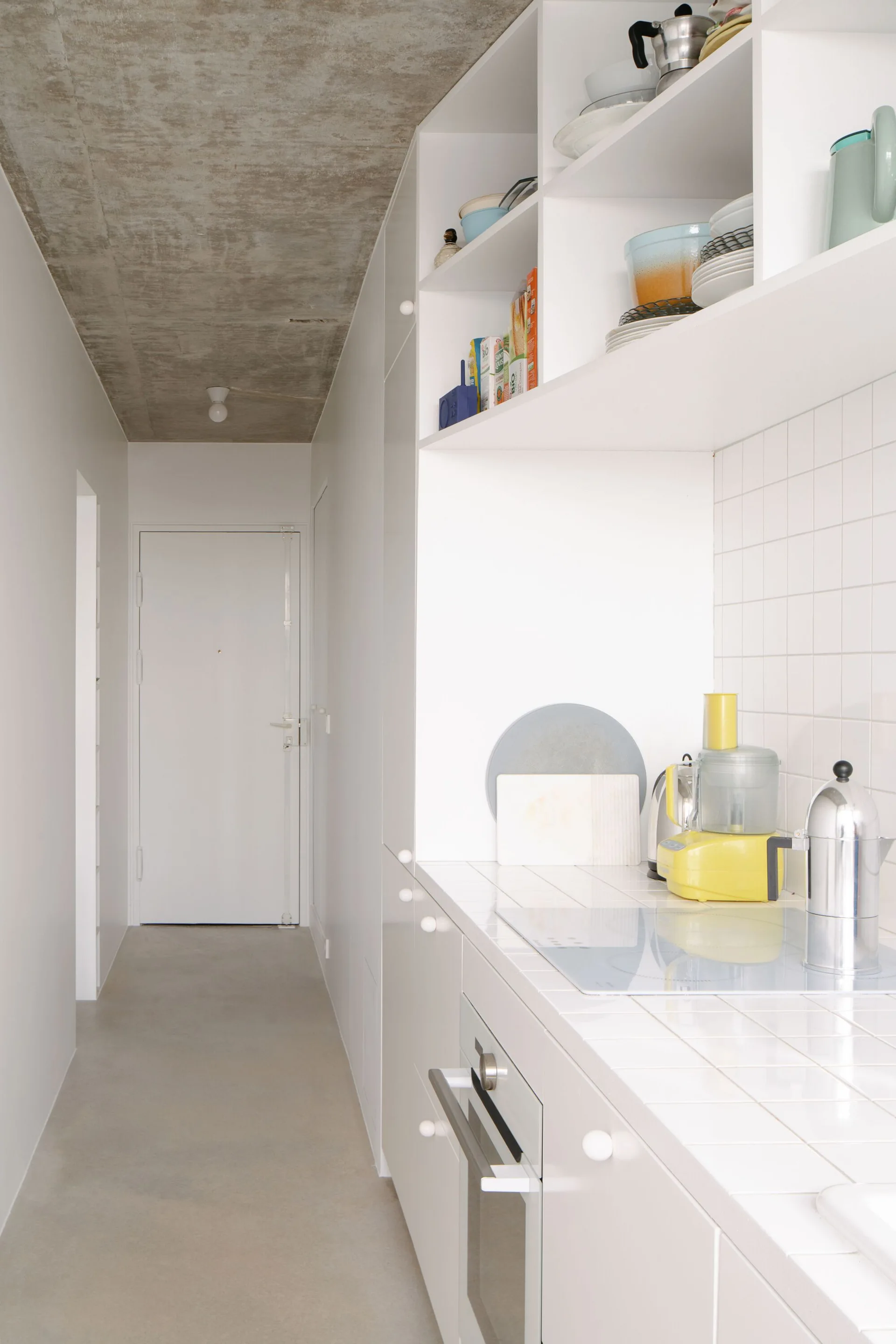 A minimalist galley kitchen features white tiled countertops and backsplash, white cabinetry, and an exposed raw concrete ceiling.