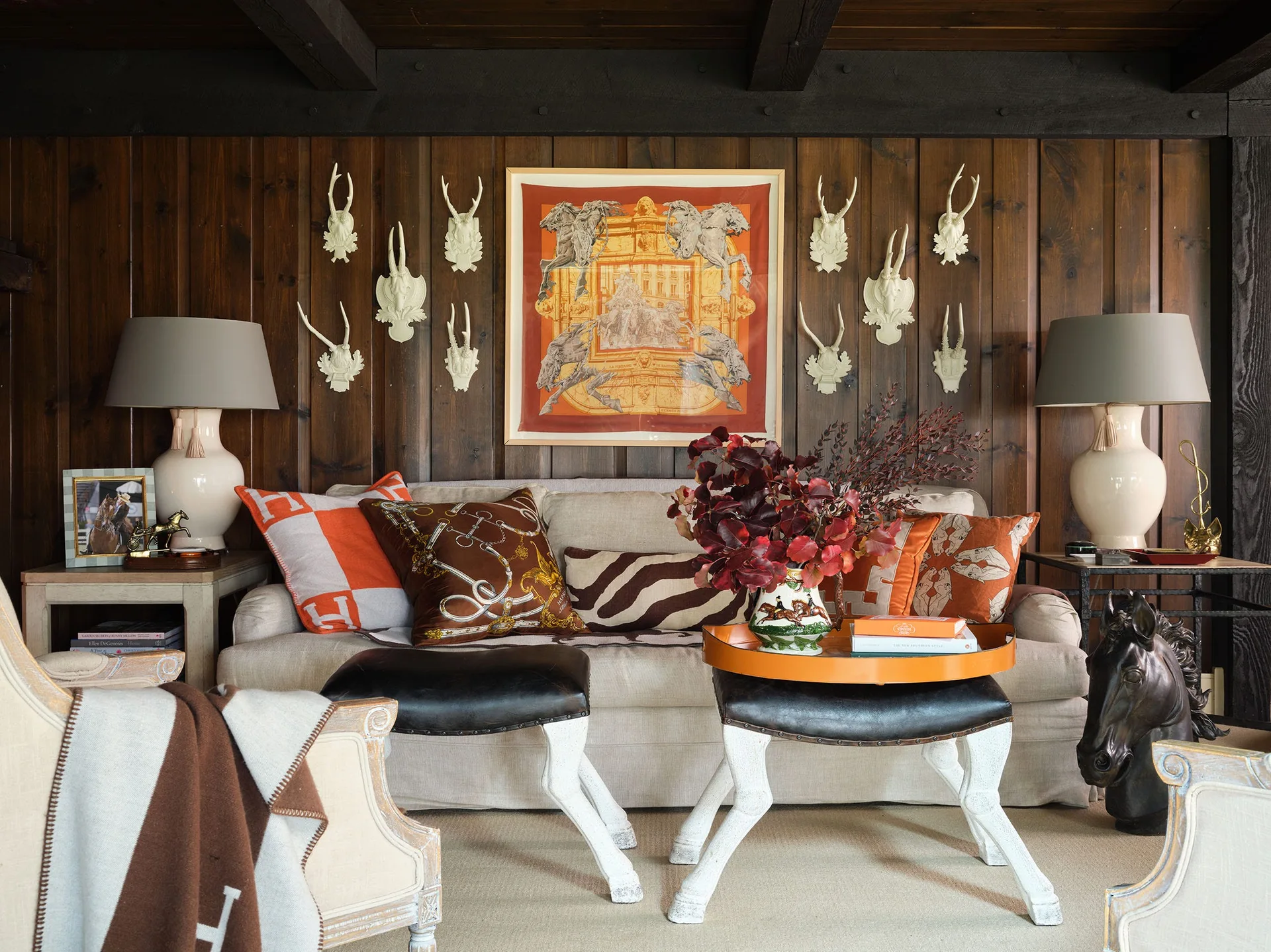 A beige sofa sits against dark wood paneled walls decorated with white antler sculptures and framed orange equestrian wall art.