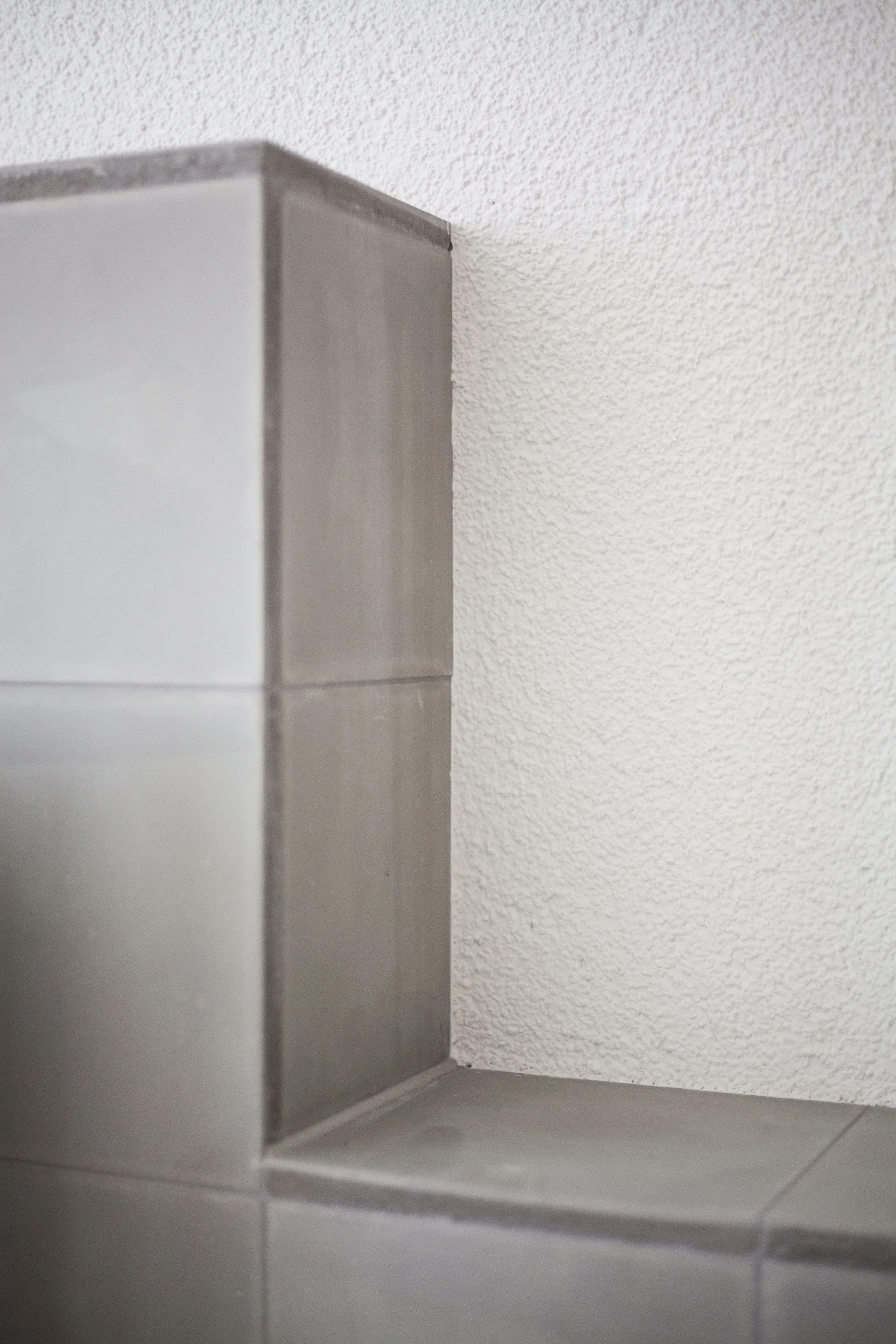 A close-up view of a minimalist corner constructed with matte gray square tiles meeting a white textured stucco plaster wall.
