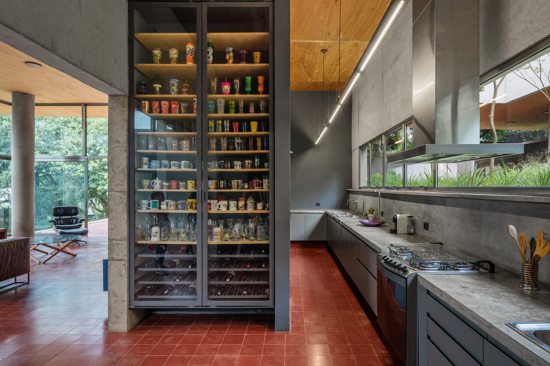 An industrial kitchen features a large glass display cabinet filled with mugs, concrete walls, a red tiled floor, and a long narrow window.
