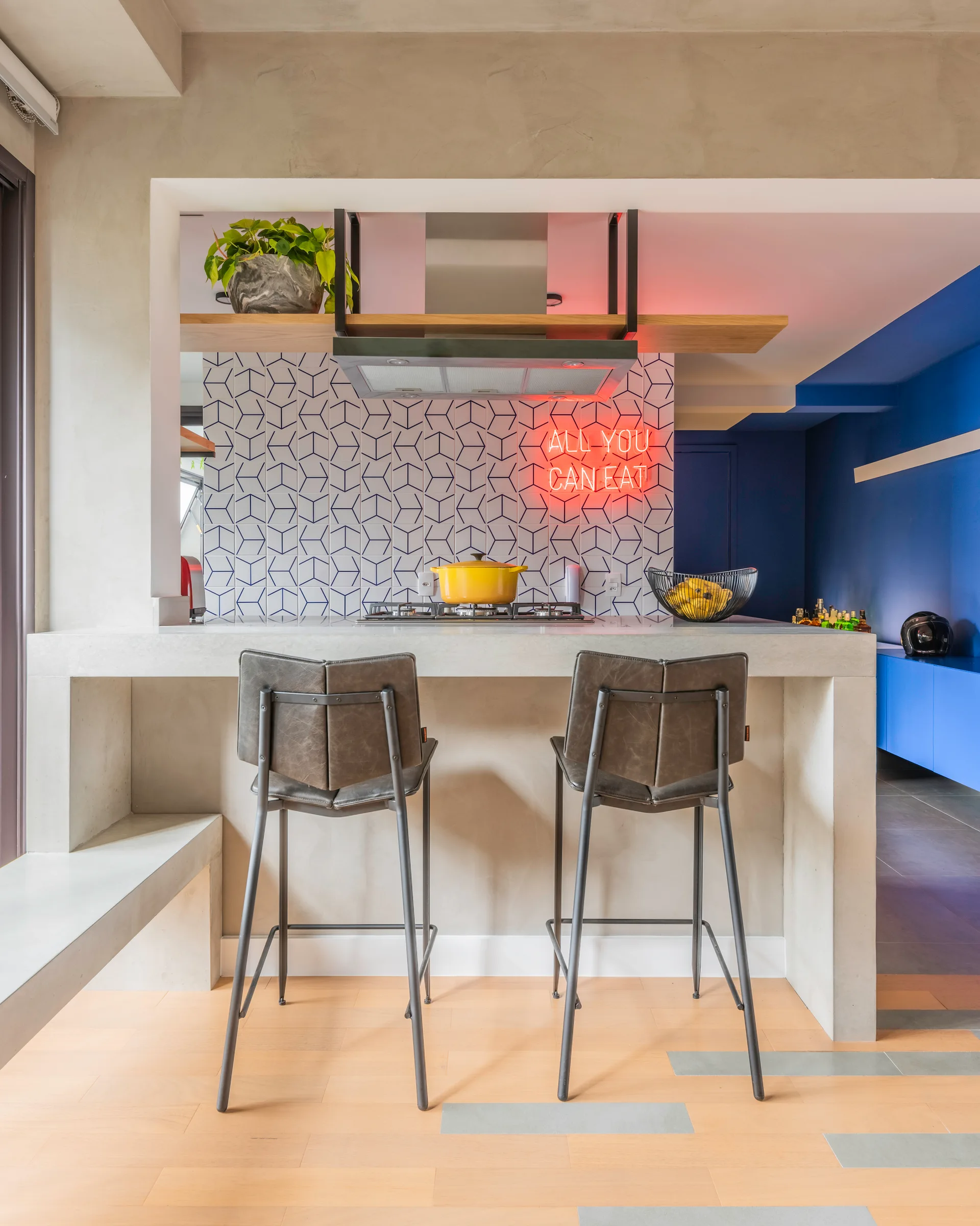 A modern kitchen features a concrete island with two gray bar stools, a blue-and-white geometric tiled backsplash, and a glowing red neon sign.