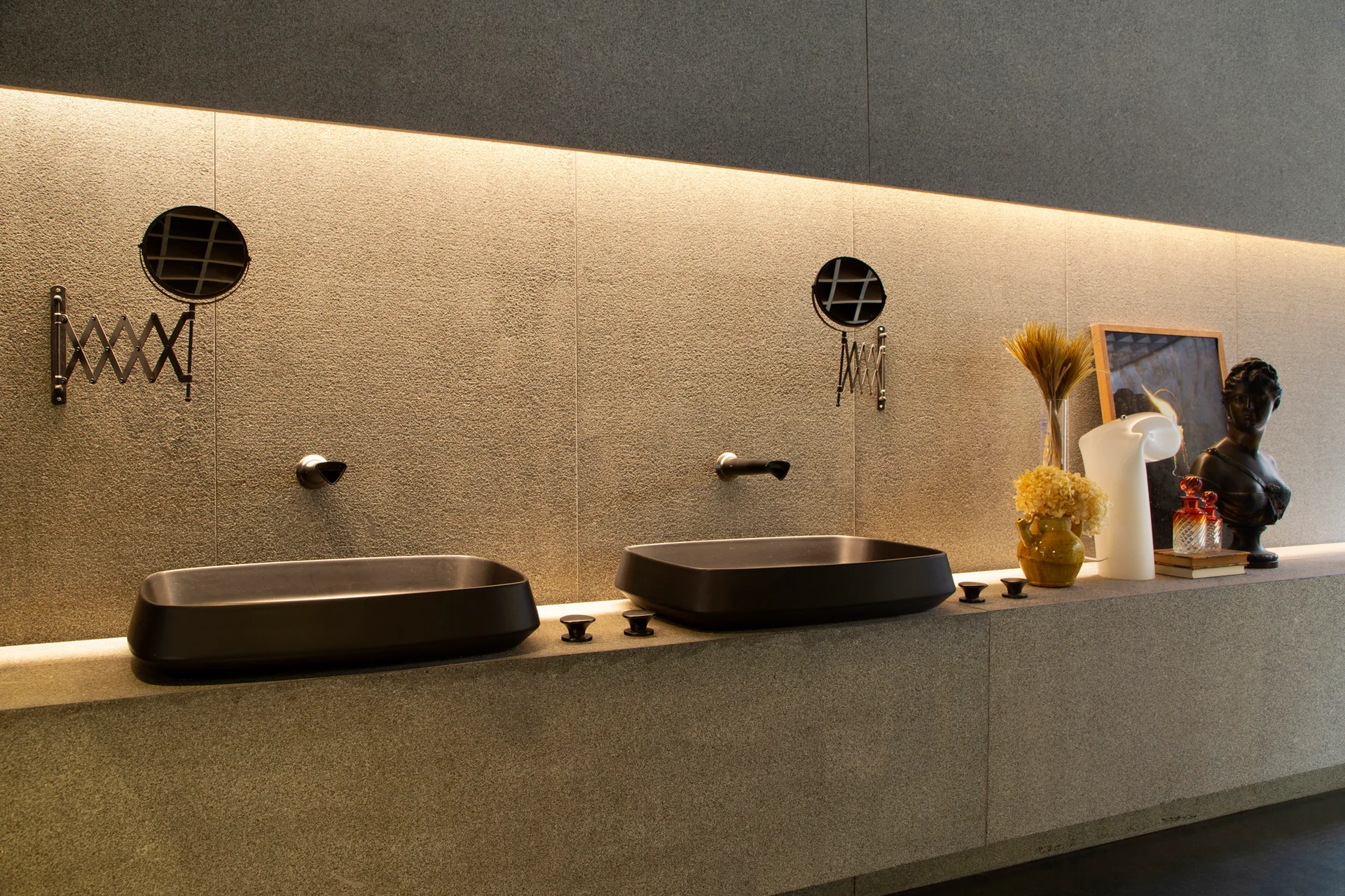 A modern gray stone bathroom vanity features two matte black vessel sinks, wall-mounted faucets, extendable mirrors, and warm recessed linear lighting.