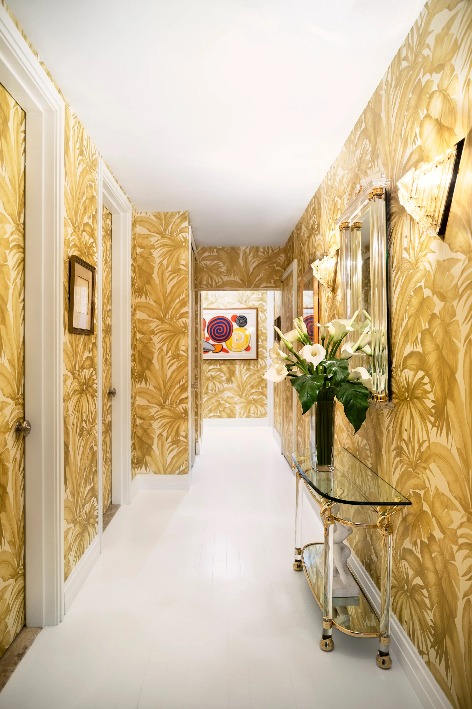 A long hallway features gold tropical leaf wallpaper, white painted floors, a glass console table with calla lilies, and a glass-framed mirror.