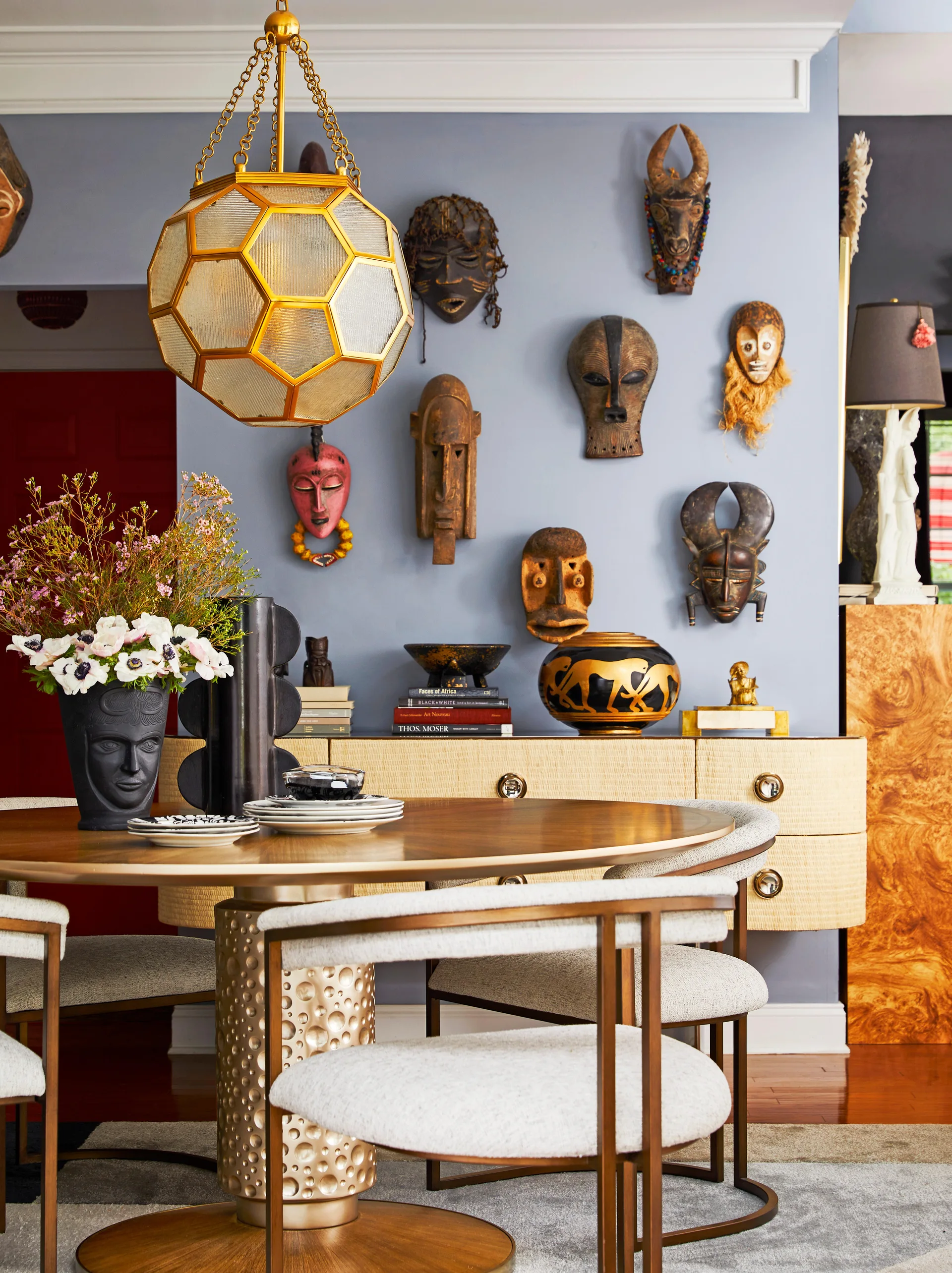 An eclectic dining room featuring a round wooden table, upholstered chairs, and a light blue wall adorned with carved wooden masks.