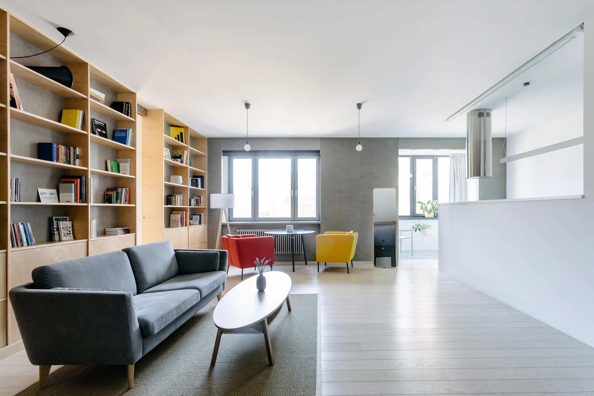 A modern open-plan living room features a large light wood bookshelf, a grey sofa, and red and yellow accent chairs near a window.