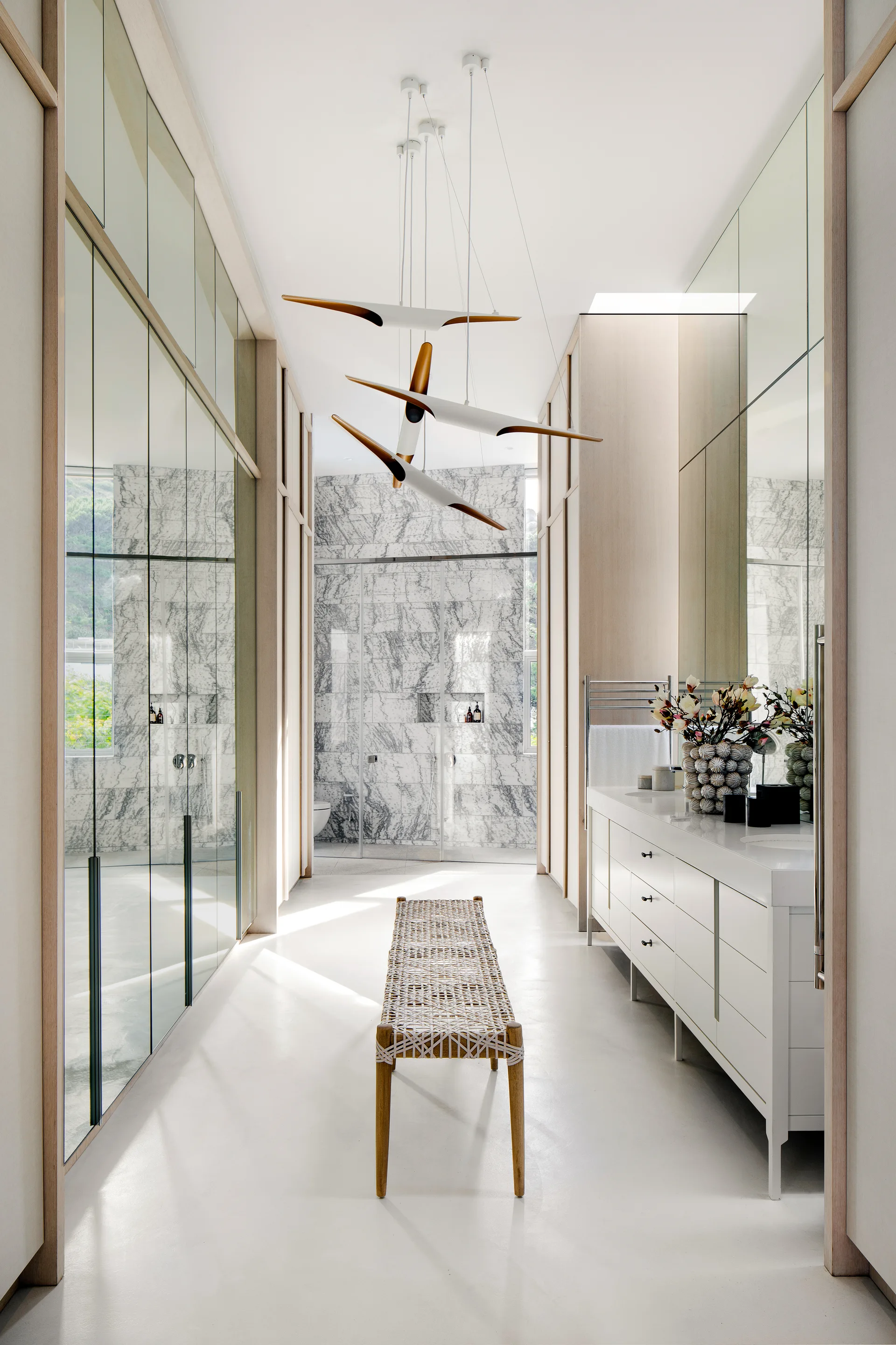 A minimalist white bathroom features a central woven wood bench, a sleek white vanity cabinet, mirrored wall panels, and a veined marble back wall.