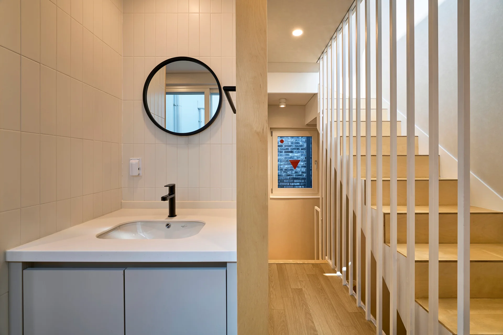 A modern powder room featuring a white tiled vanity with a round black mirror, situated next to a light wood staircase with white vertical railings.