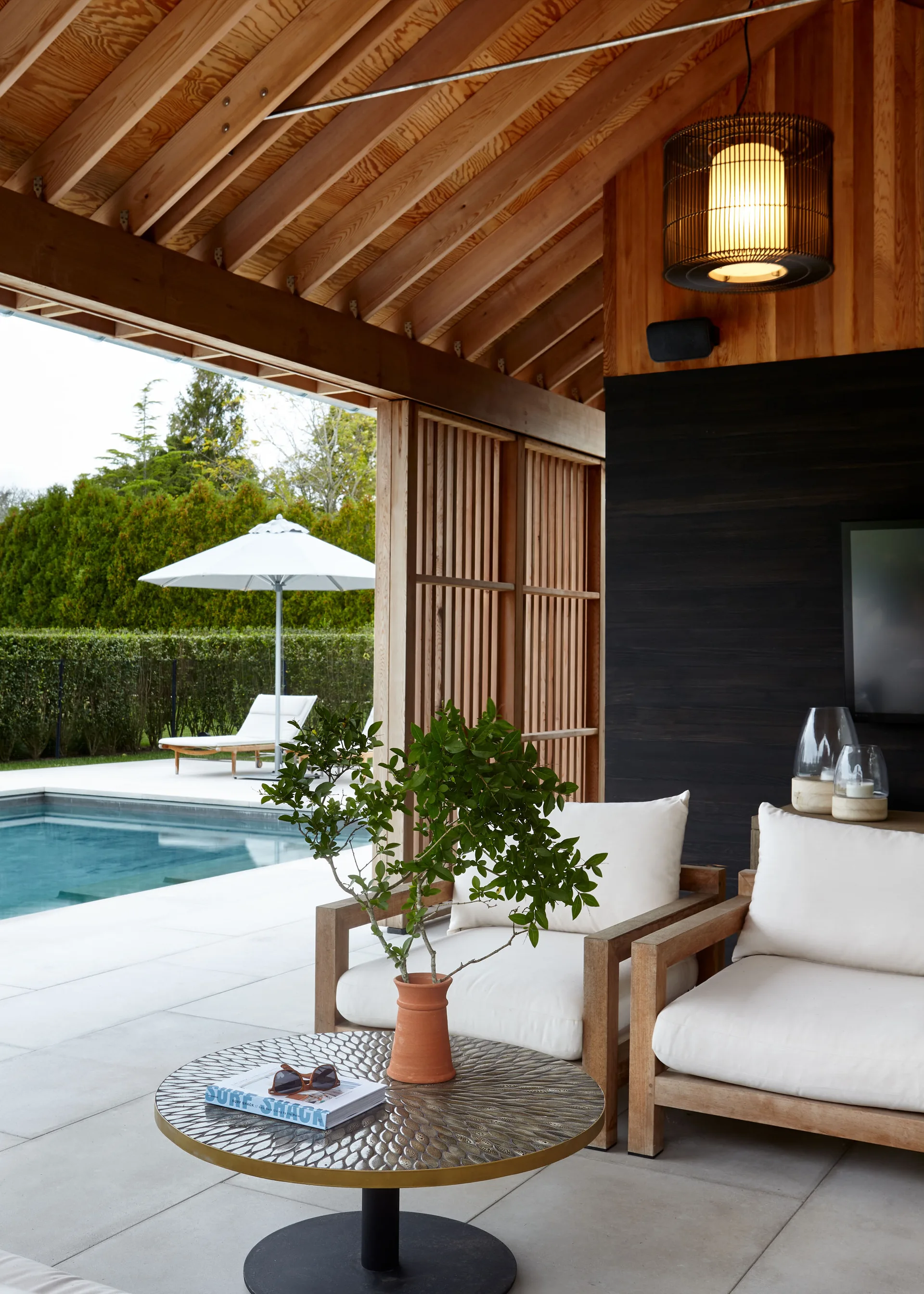 A covered patio with wood rafters and white armchairs looks out toward a swimming pool with a white umbrella.