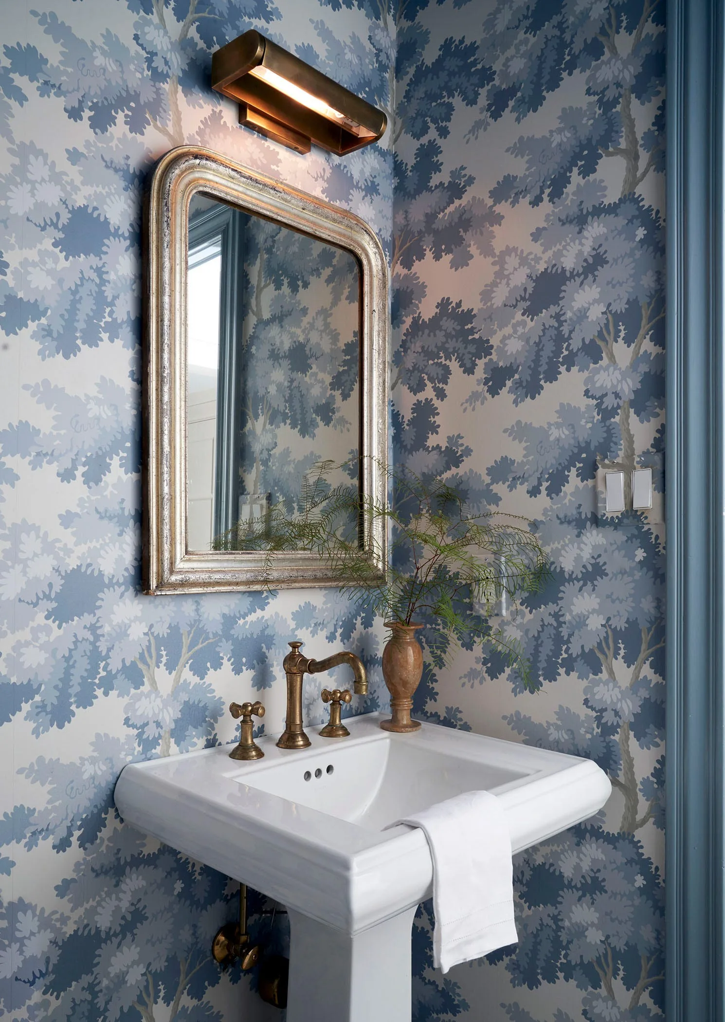A powder room features blue tree-patterned wallpaper, a white pedestal sink with brass fixtures, a silver-framed mirror, and a brass wall sconce.