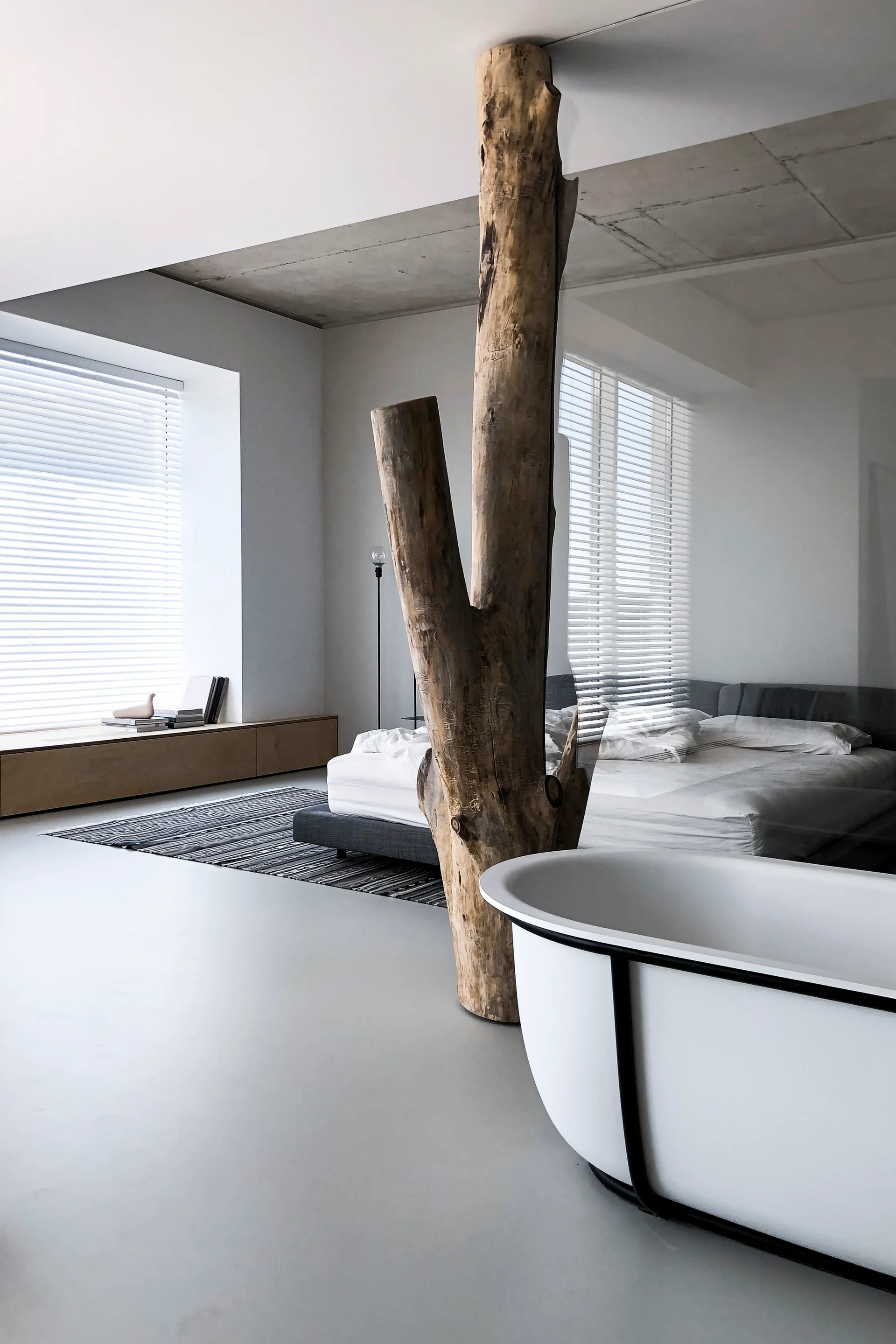 A minimalist bedroom features a large raw tree trunk column integrated into a glass wall beside a white freestanding bathtub.