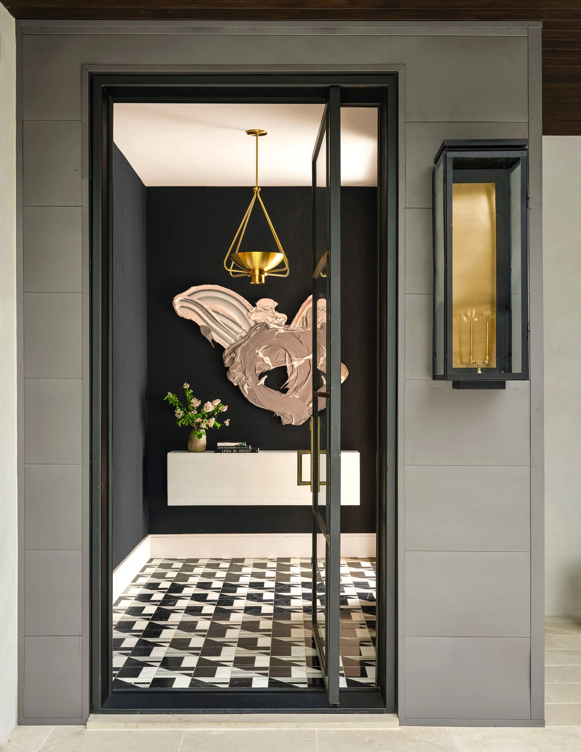 An entryway seen through an open glass door features textured black walls, a white floating console, a geometric tiled floor, and a gold pendant light.