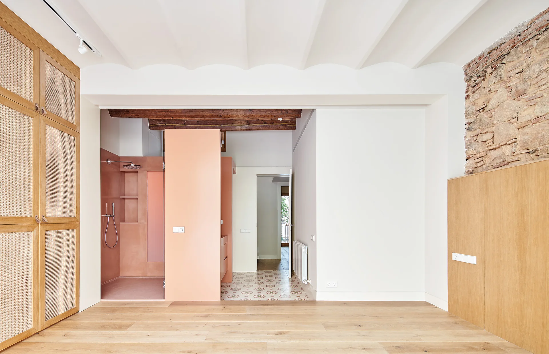 An open bedroom space features light wood floors, a large cane cabinet, a pink plaster shower enclosure, and a partial exposed stone wall.