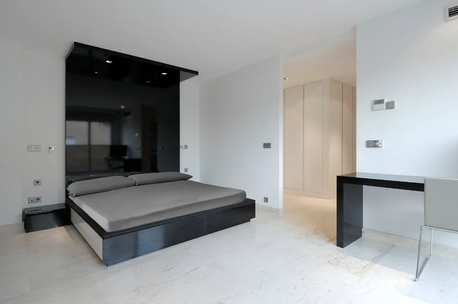 A modern bedroom features a low platform bed set against a tall glossy black headboard panel, with light stone flooring and a black console table.