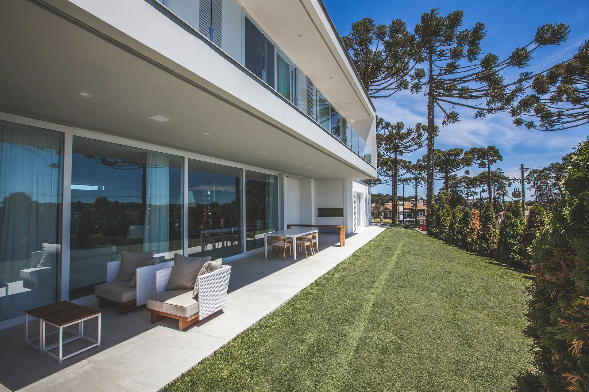 A modern concrete patio features white wicker armchairs, a dining set, and large sliding glass doors leading into the home next to a grassy lawn.