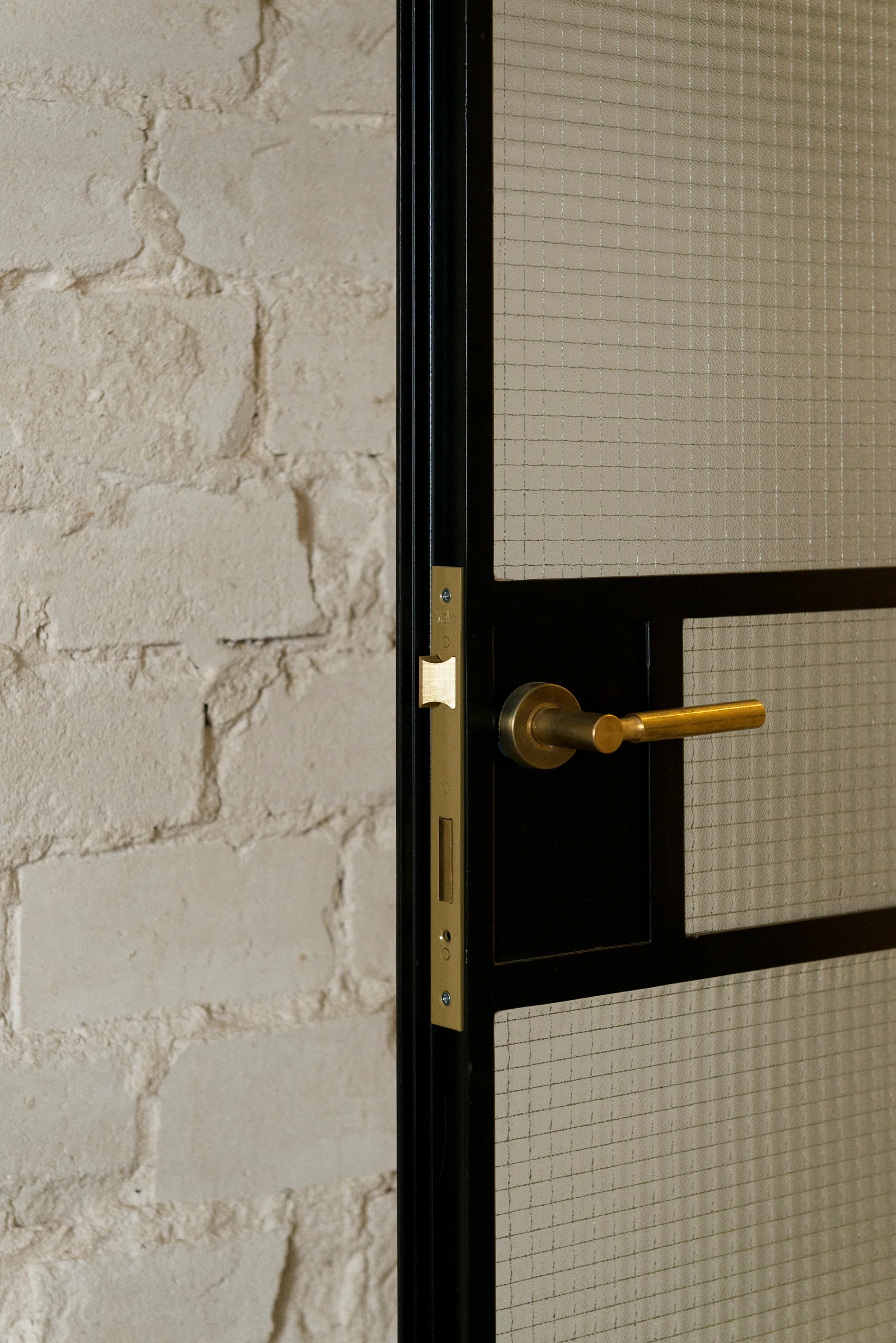 A close-up of a black metal-framed door with wire glass panels and a brass handle set against a painted brick wall.