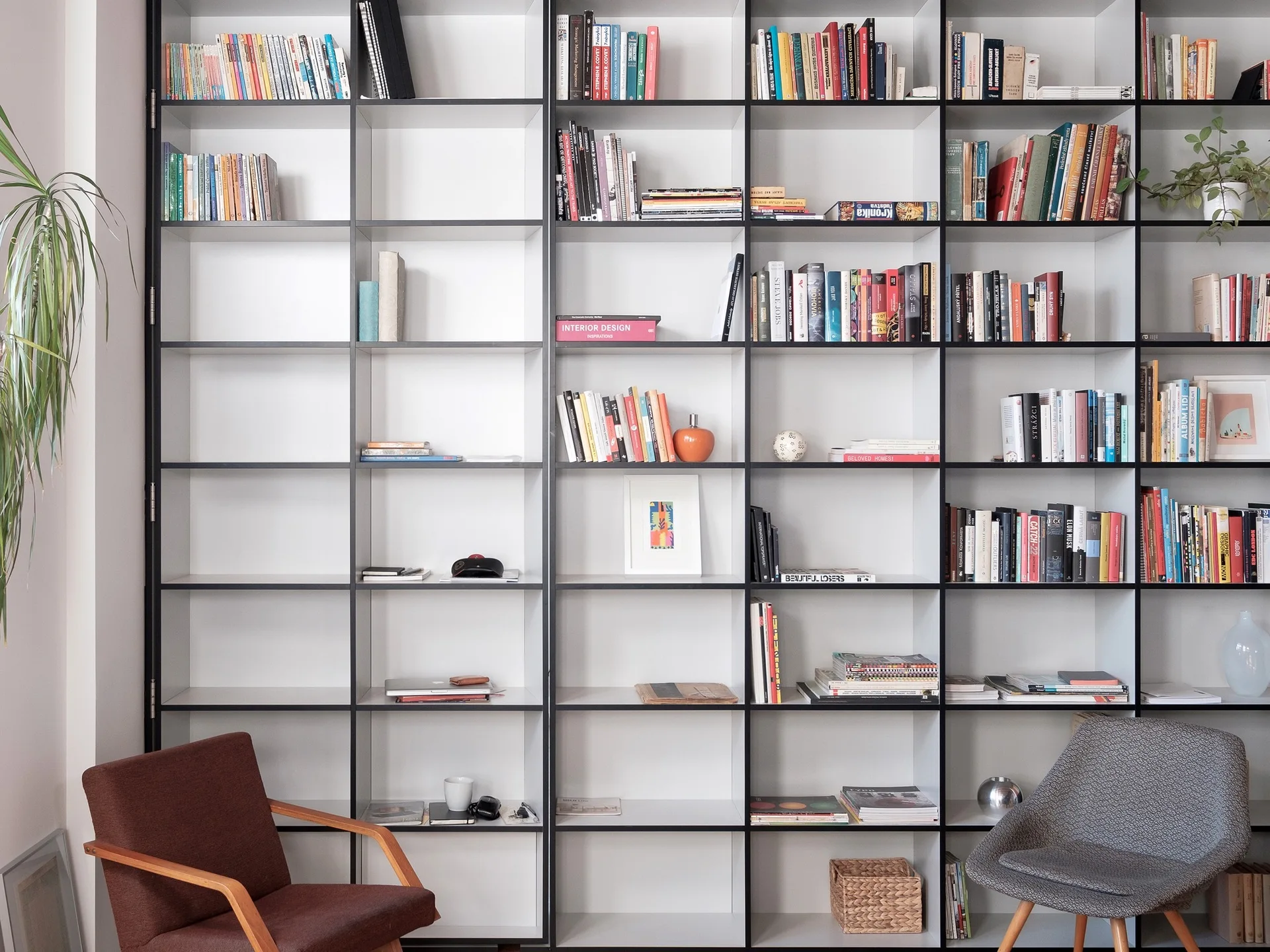 A floor-to-ceiling white grid bookshelf filled with books and decor sits behind a brown wood armchair and a grey fabric armchair.