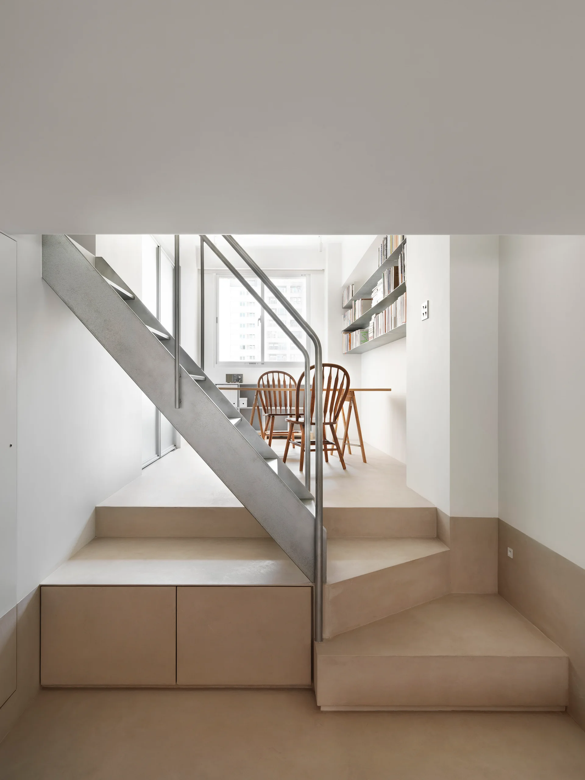 A minimalist interior features a metal staircase with built-in storage leading up to a bright dining space with wooden chairs.