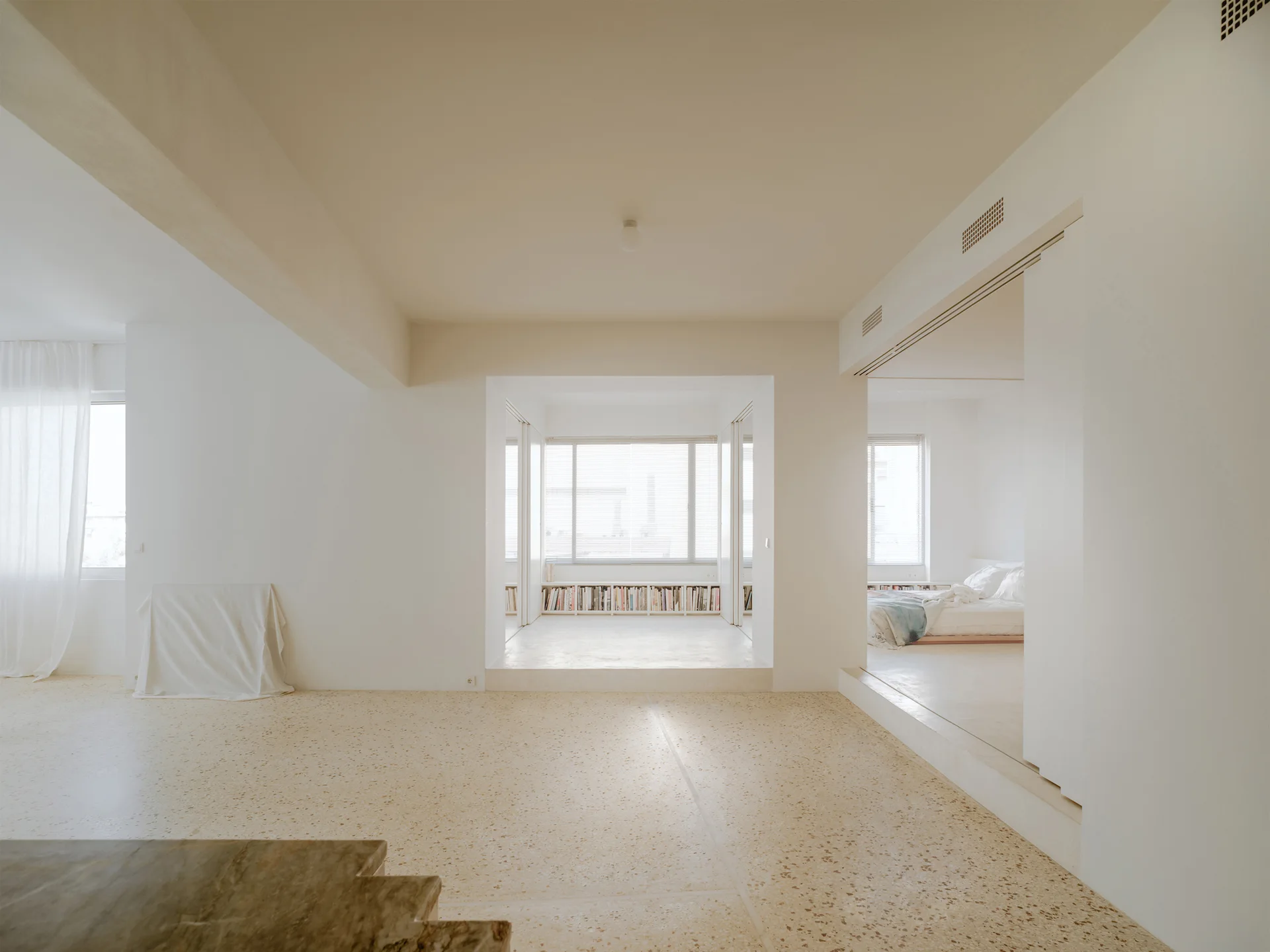 A minimalist open-plan interior features beige terrazzo flooring, smooth plaster walls, and raised platform entryways into adjacent rooms.