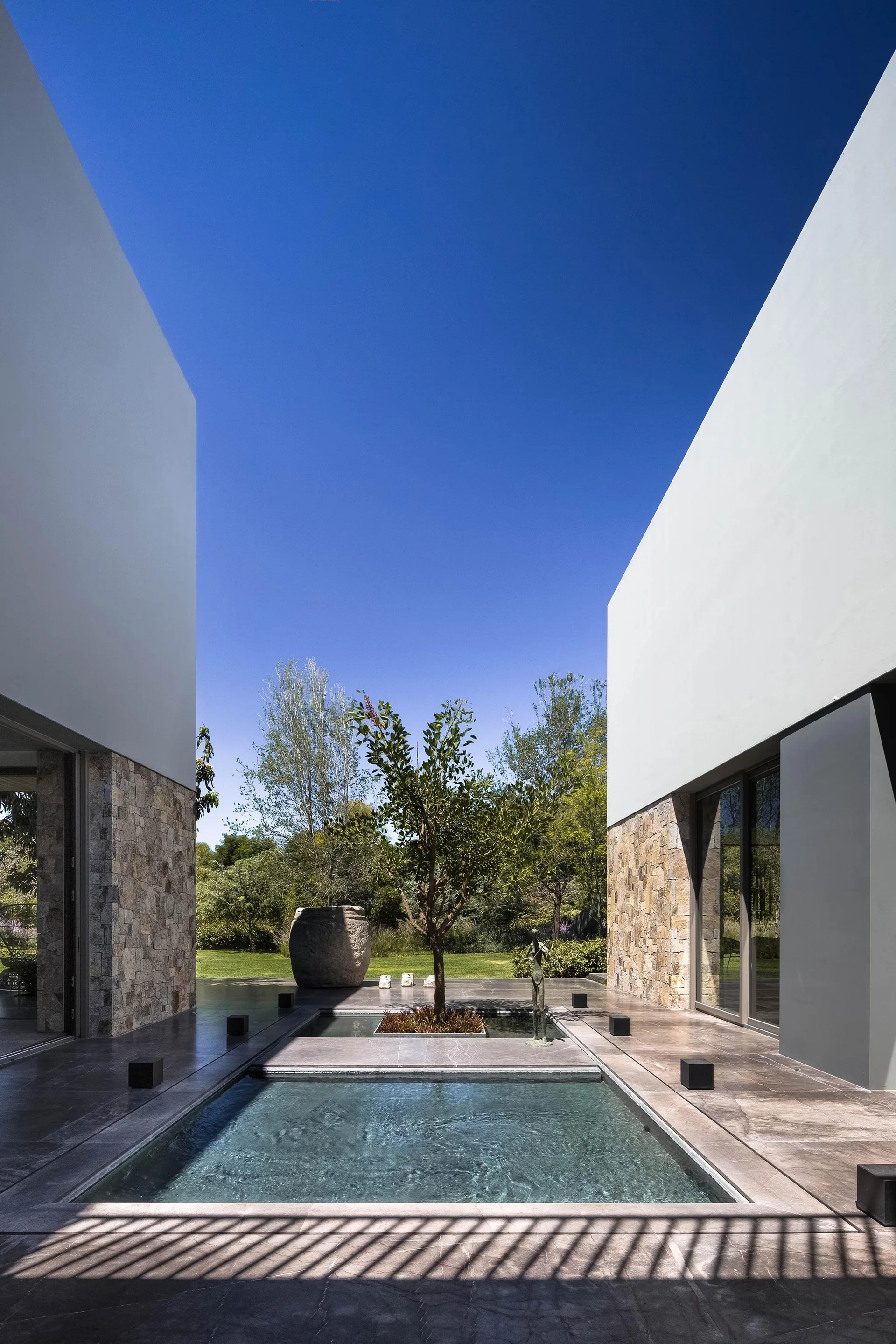 A modern outdoor courtyard featuring a central rectangular water pool, grey stone tile flooring, a small tree, and stucco walls with stone cladding.